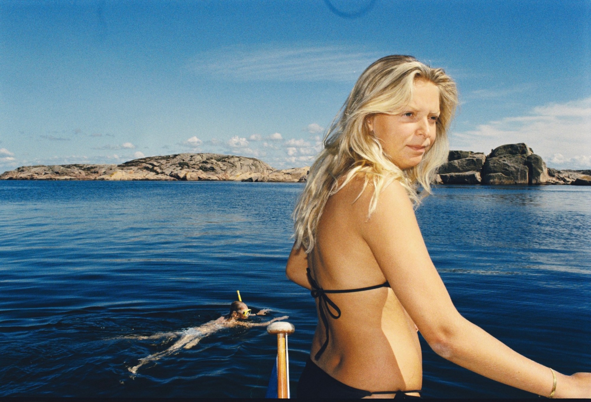 a man swimming in the water in sweden while a blonde woman stands in the foreground