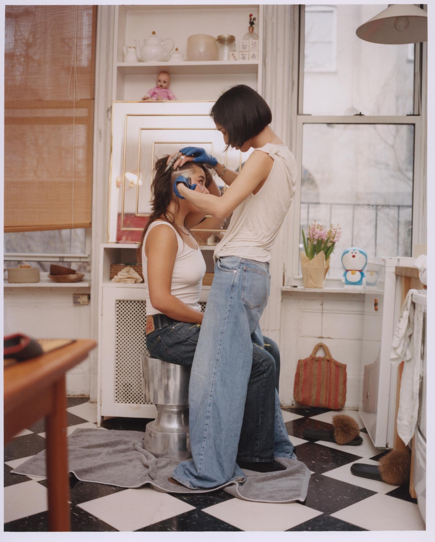 two women in a kitchen with black and white tiles, one cutting the other's hair