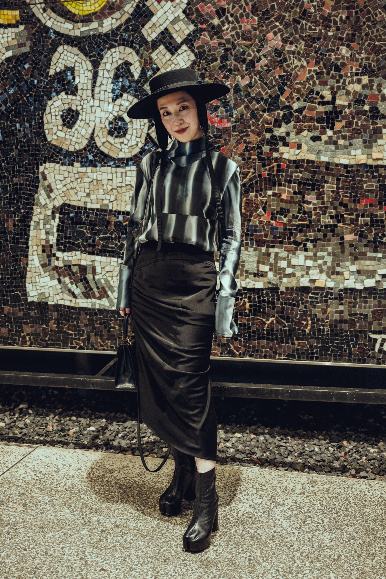 a japanese woman stands in front of a wall mosaic wearing tabi boots, a rouched skirt and a monochrome shirt. there's a large-rimmed hat resting on her head