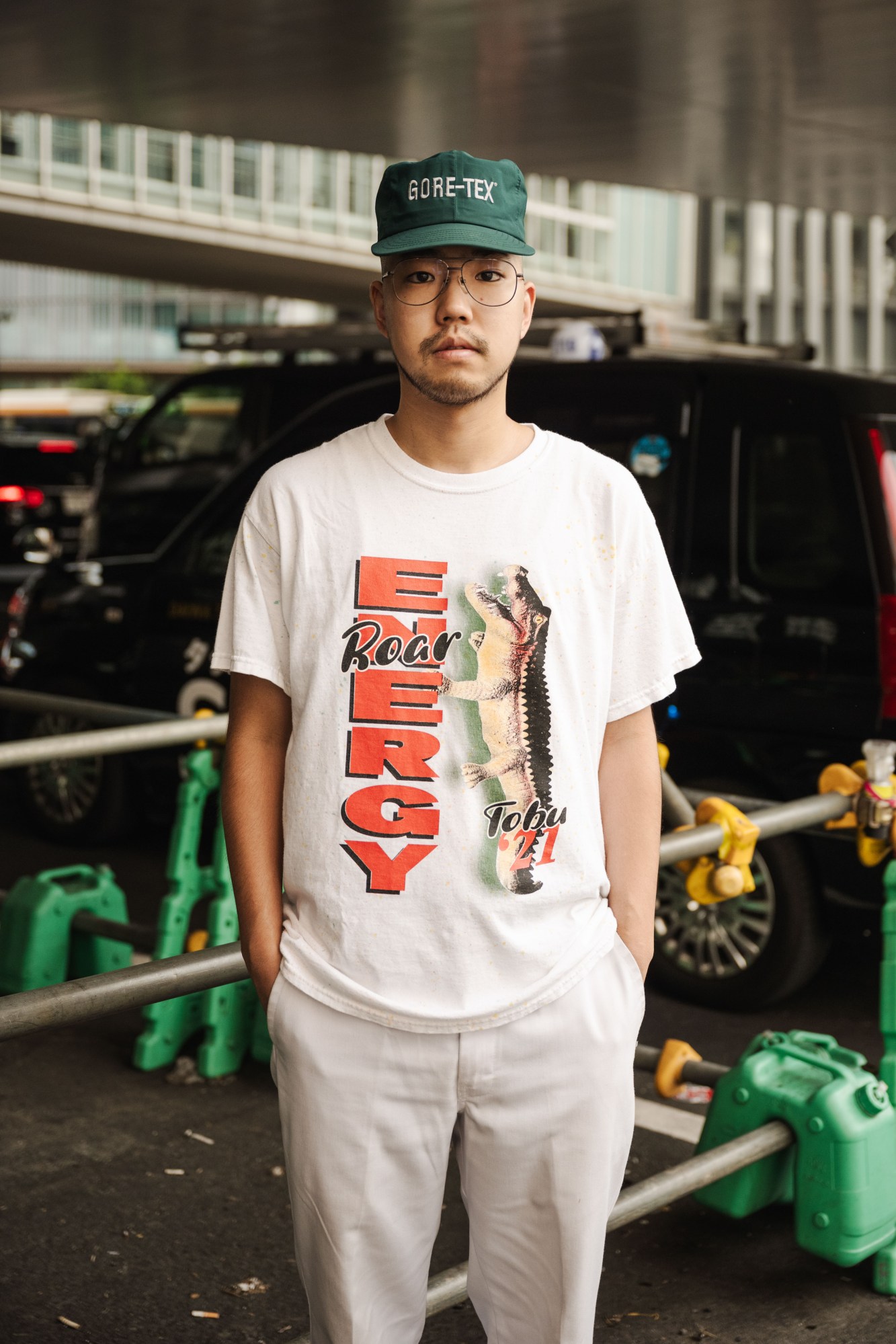 a japanese man stands with his hands in his white trouser pockets. his T-shirt has a crocodile on it with the words ENERGY, and he's wearing a cap with wire-rimmed glasses