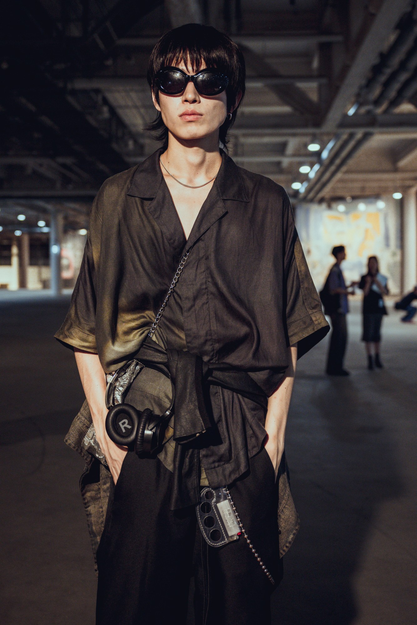 a young japanese man wearing baggy black trousers and an oversized shirt with dark sunglasses. there are a pair of overear headphones hanging from his bag strap