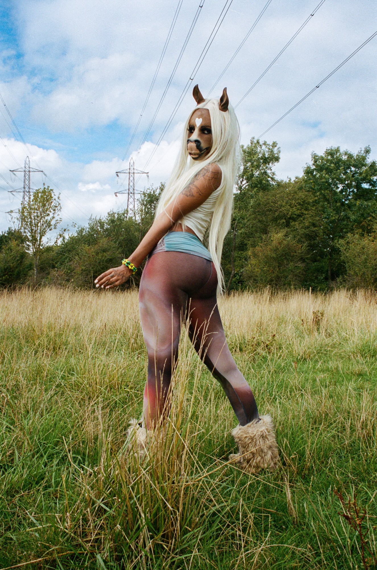 the DJ horsegiirL stands facing away from the camera in a field, she looks back over her shoulder to camera