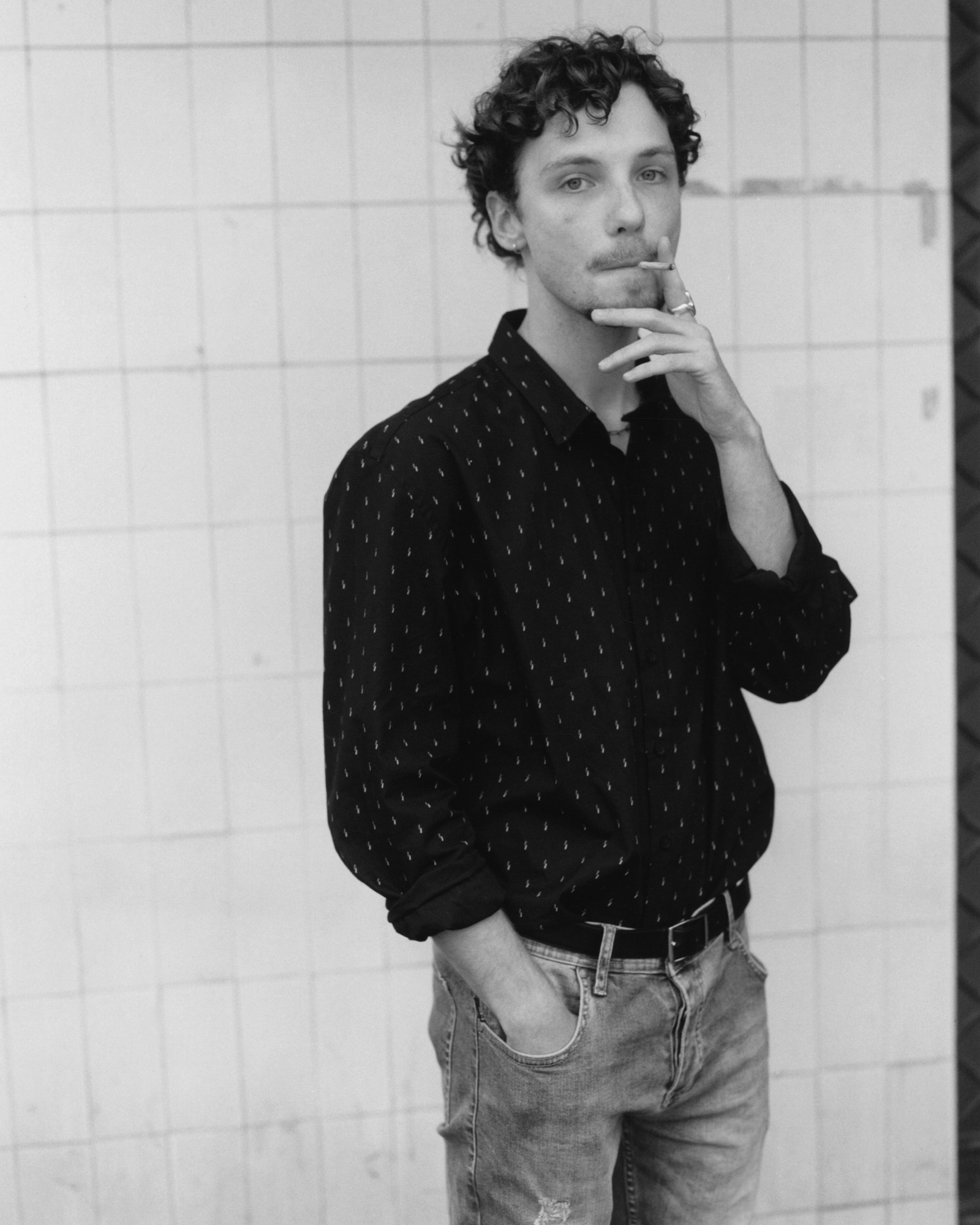 the actor jeremy gillet smoking a cigarette as he stands, hand in jean pocket, against an outside tiled wall