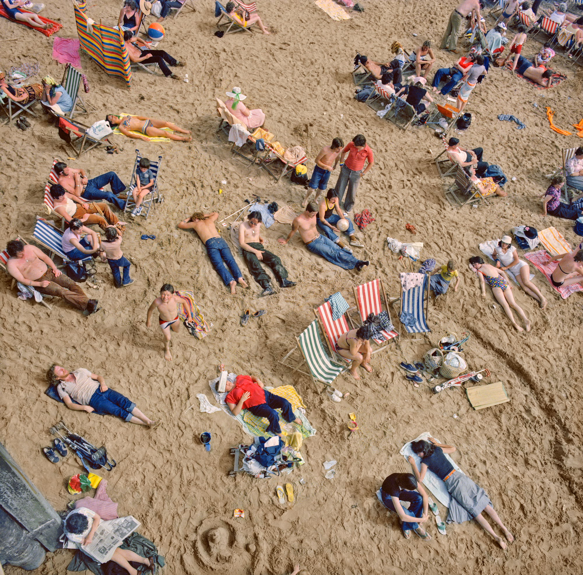 a charles traub birds eye shot of a beach looking down on sand, deckchairs, two dozen bodies wearing swimwear