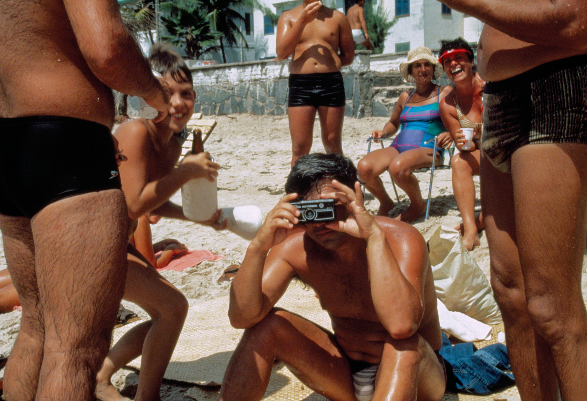 a man on an italian beach in the 1980s surrounded by other people takes a photo