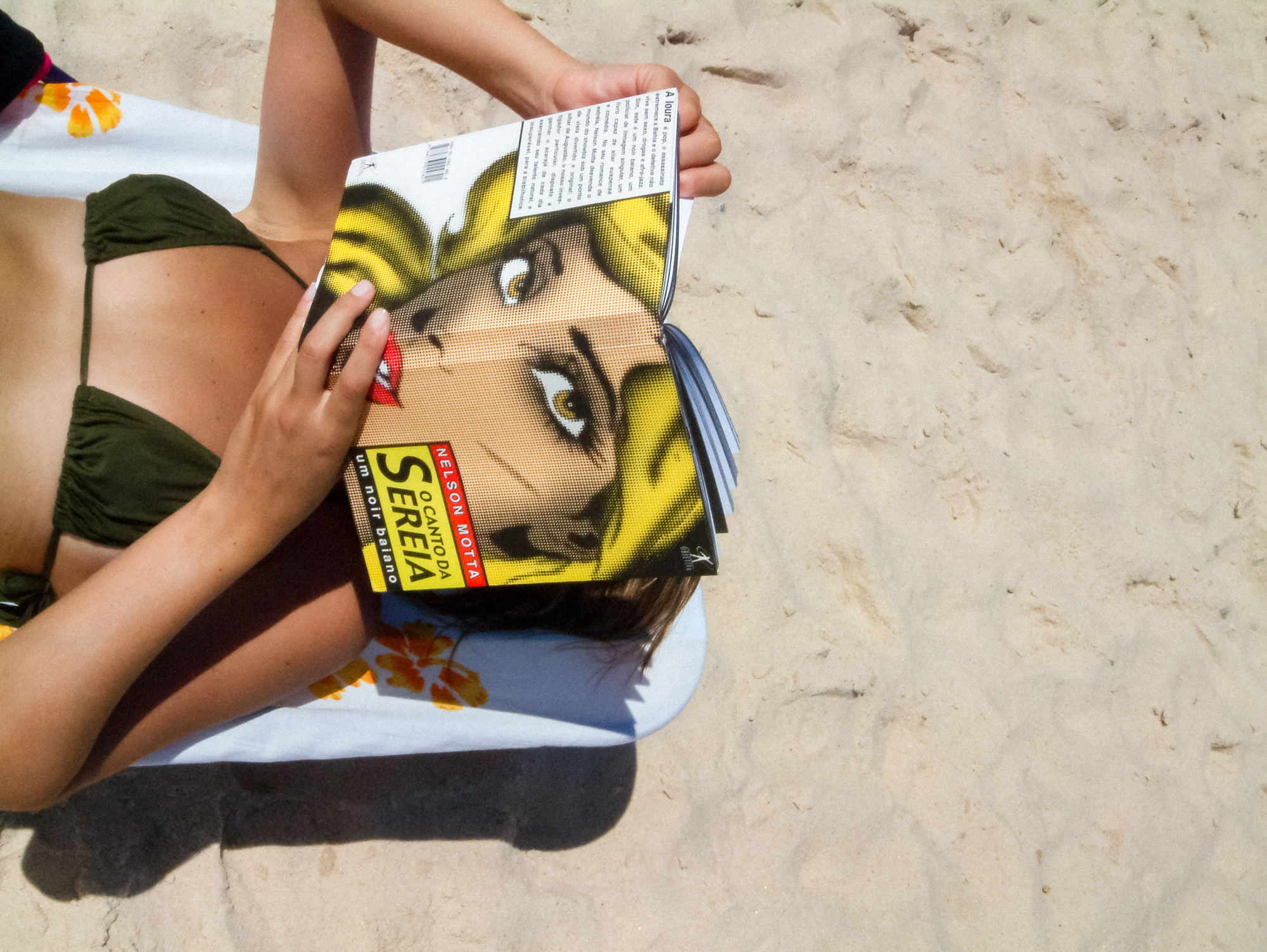 a woman on a beach in a bikini reads a book with the face of a woman on the cover