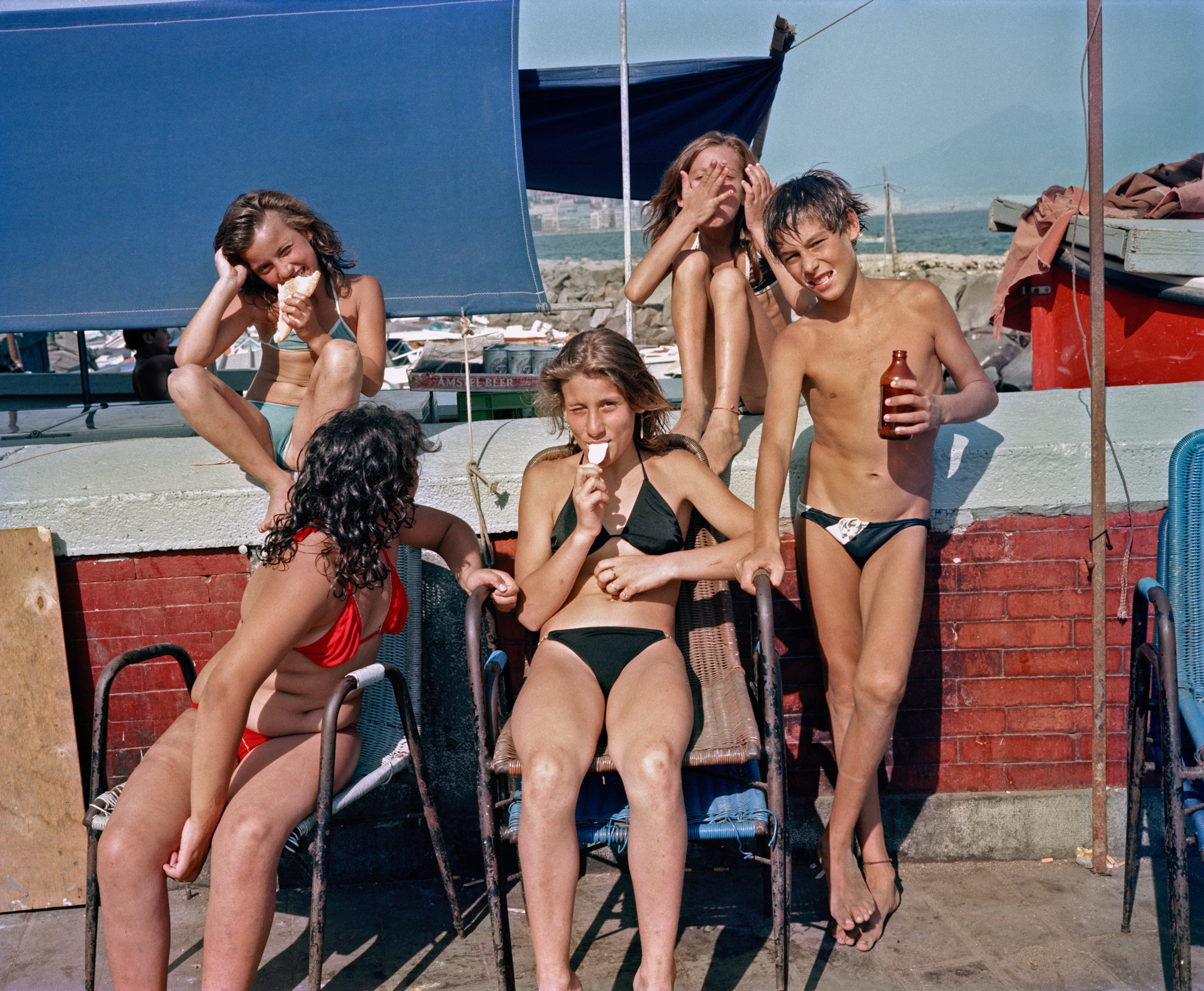 a group of teenagers in naples sit on chairs by the beach wearing swimwear, eating ice cream and drinking