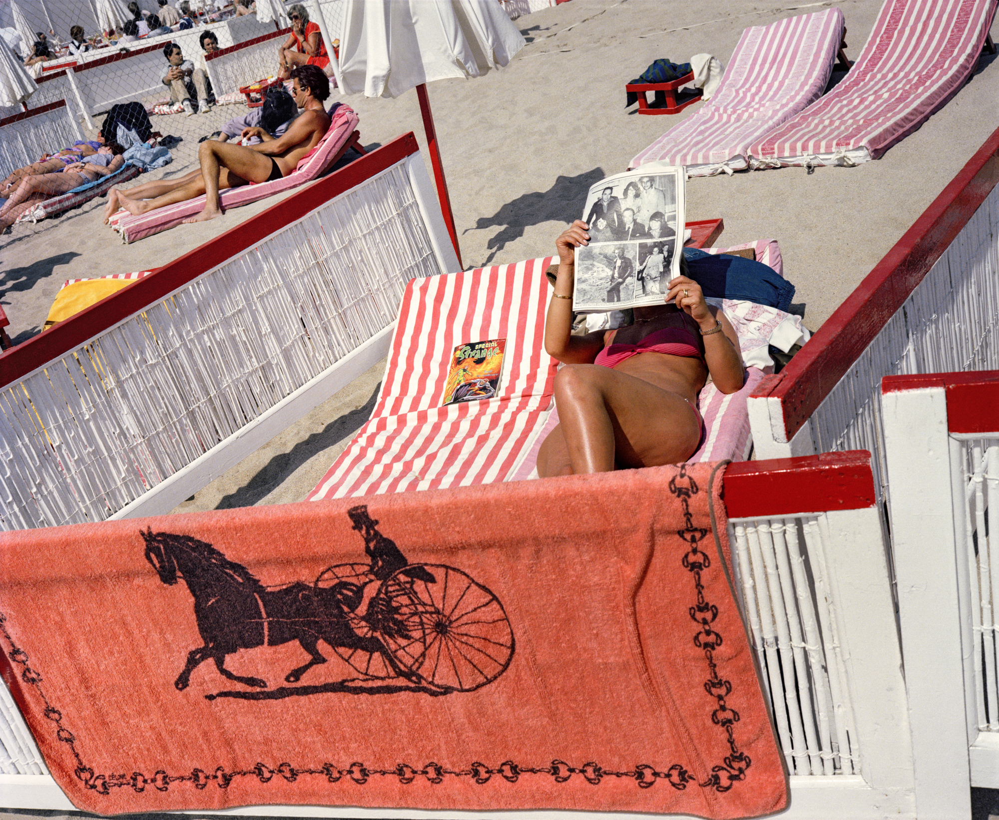 a woman lies on the beach surrounded by windbreakers, wearing a bikini and reading a newspaper