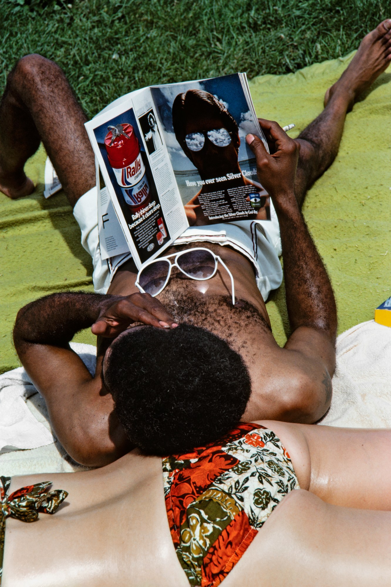 a man rests his head on a sunbathing girl's back while reading a magazine