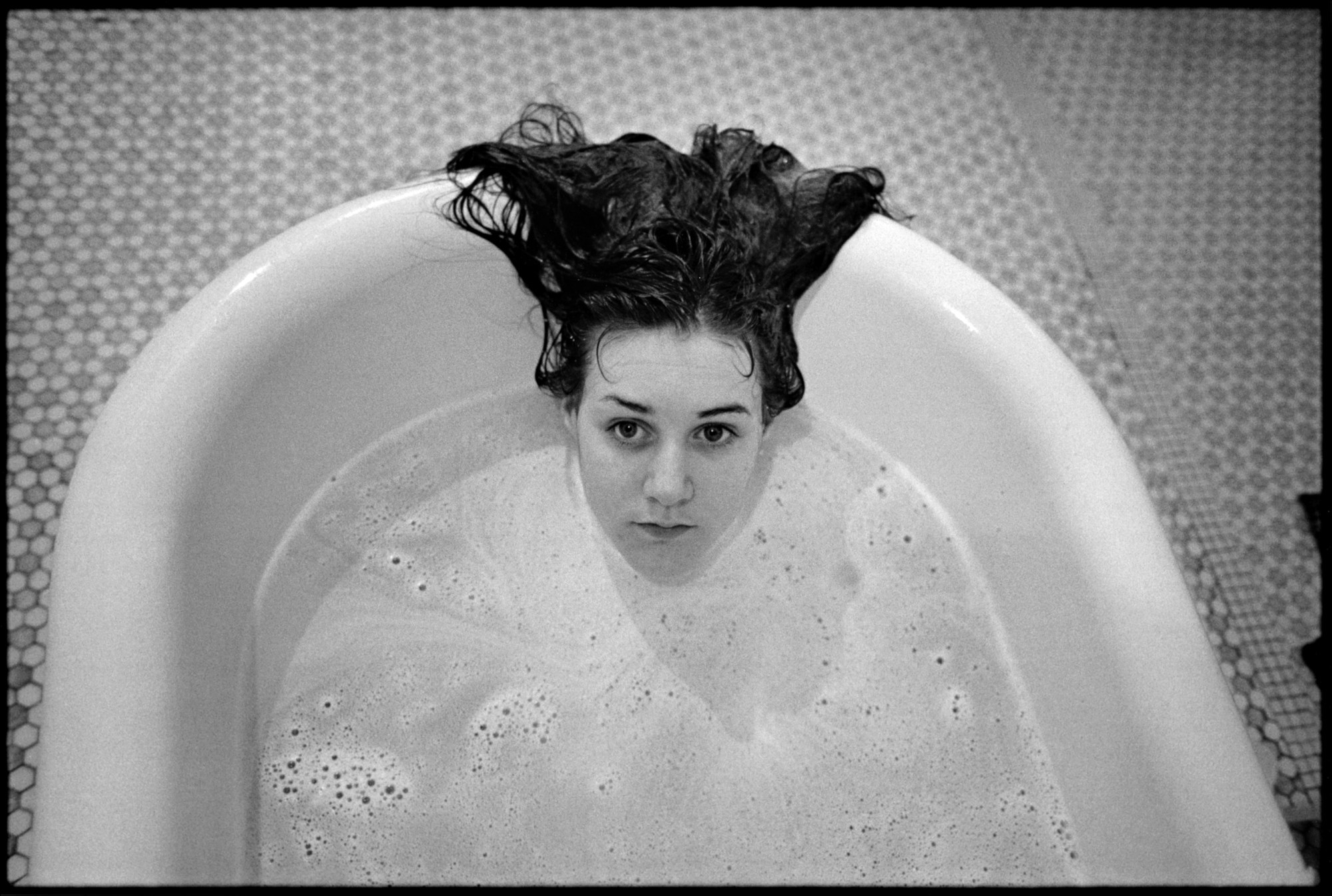 a b&w portrait of a young person almost fully submerged in a roll-top bubble bath; their face visible, with long messy hair resting back over the tub