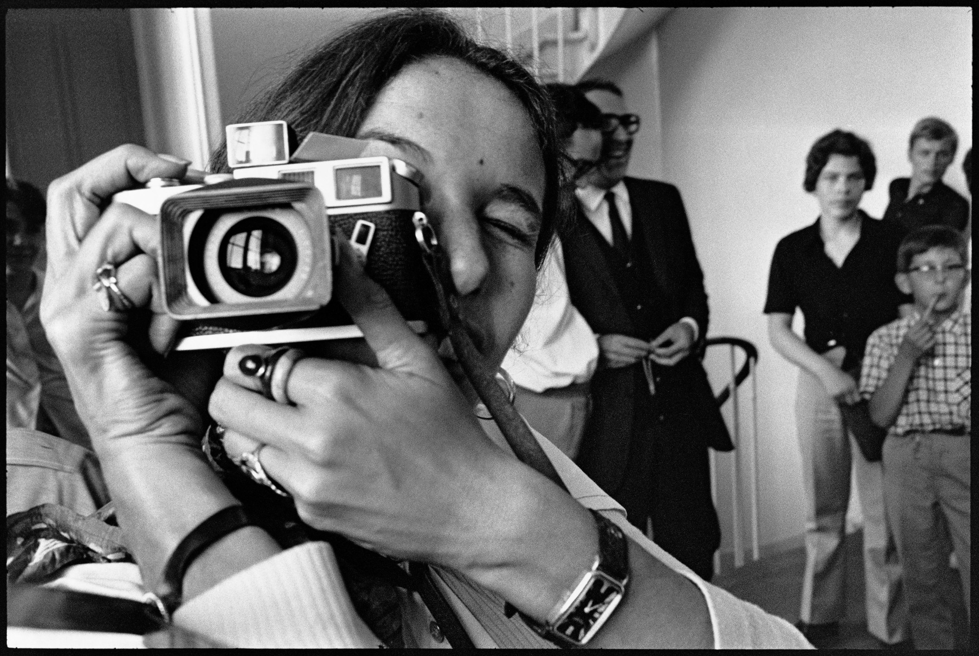 a close up of a woman with dark hair taking a photograph; a family stand around in the background