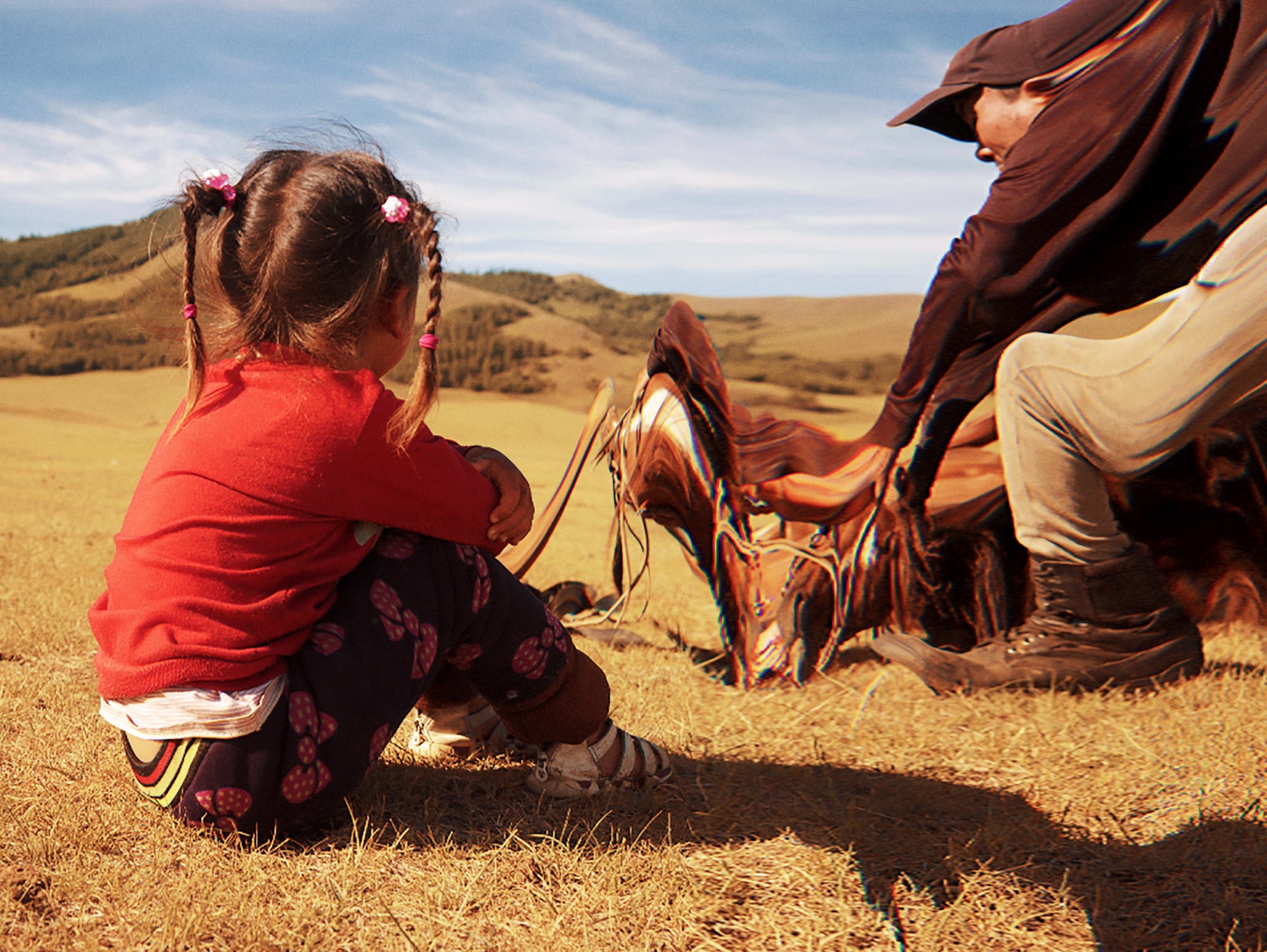 a mongolian child with their hair in braids sits in the countryside next to a herder and their horse