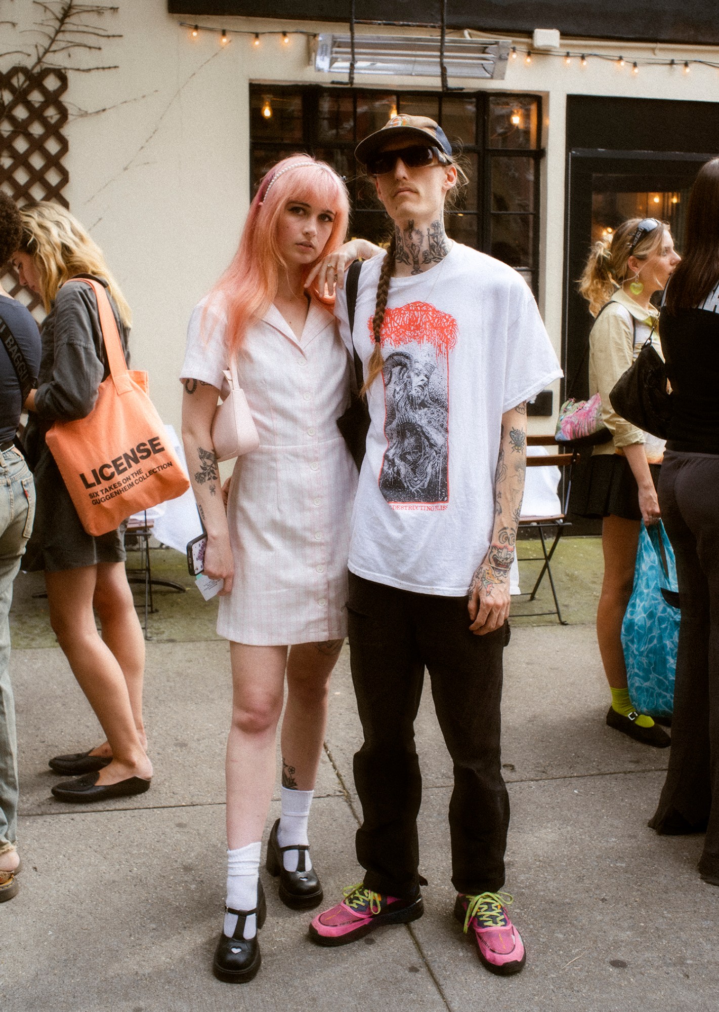 ashley markle photo of two sofia coppola fans on the streets of new york, one wearing a light gingham dress and black cute dress shoes, with pink hair. a man next to her is tattooed wearing pink sneakers, black jeans and a white t shirt.