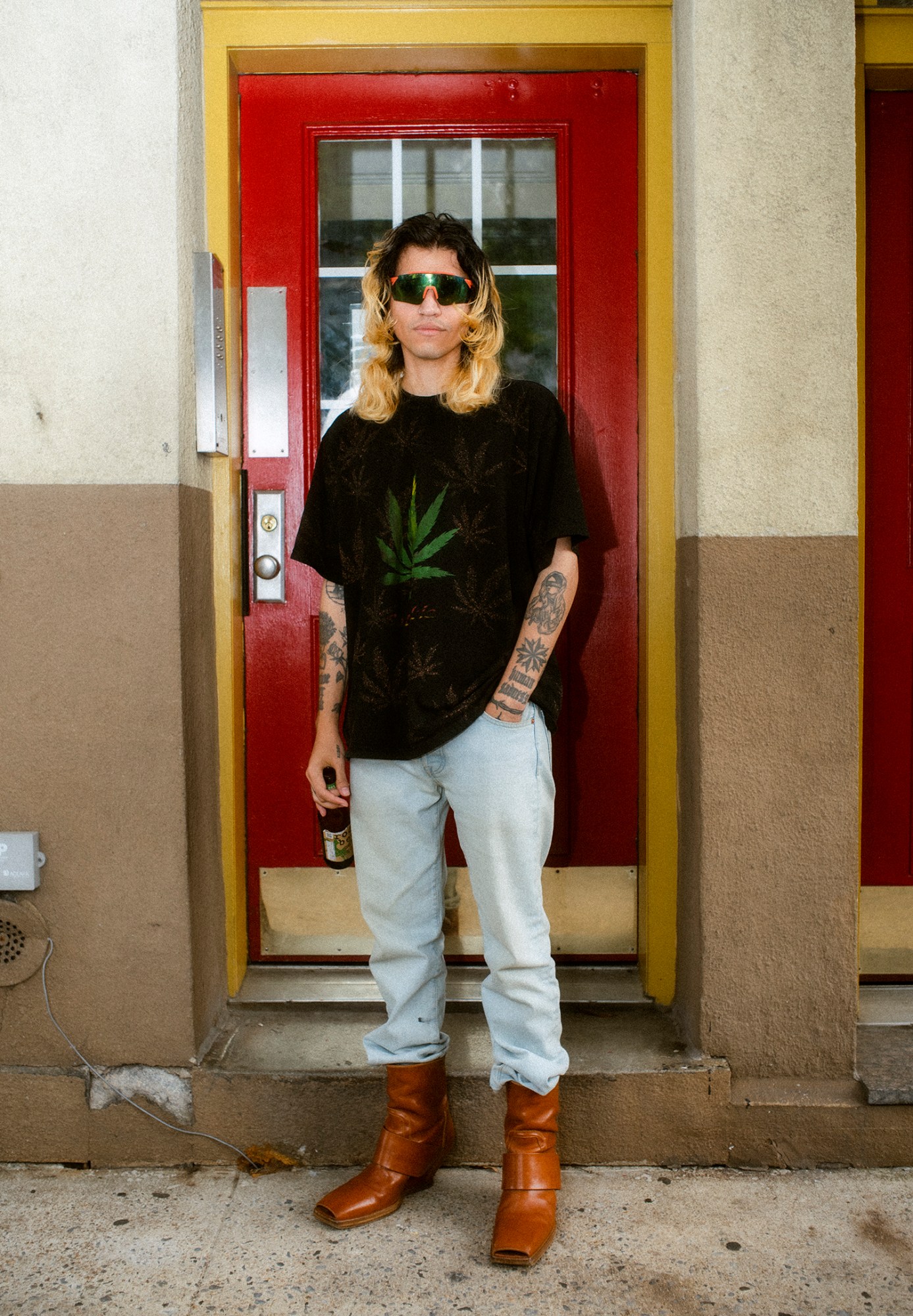 ashley markle photo of a sofia coppola fan on the streets of new york wearing tan cowboy boots, blue denim tucked in and a black t-shirt with marijuana leaves on it. they're carrying a beer and have long hair.