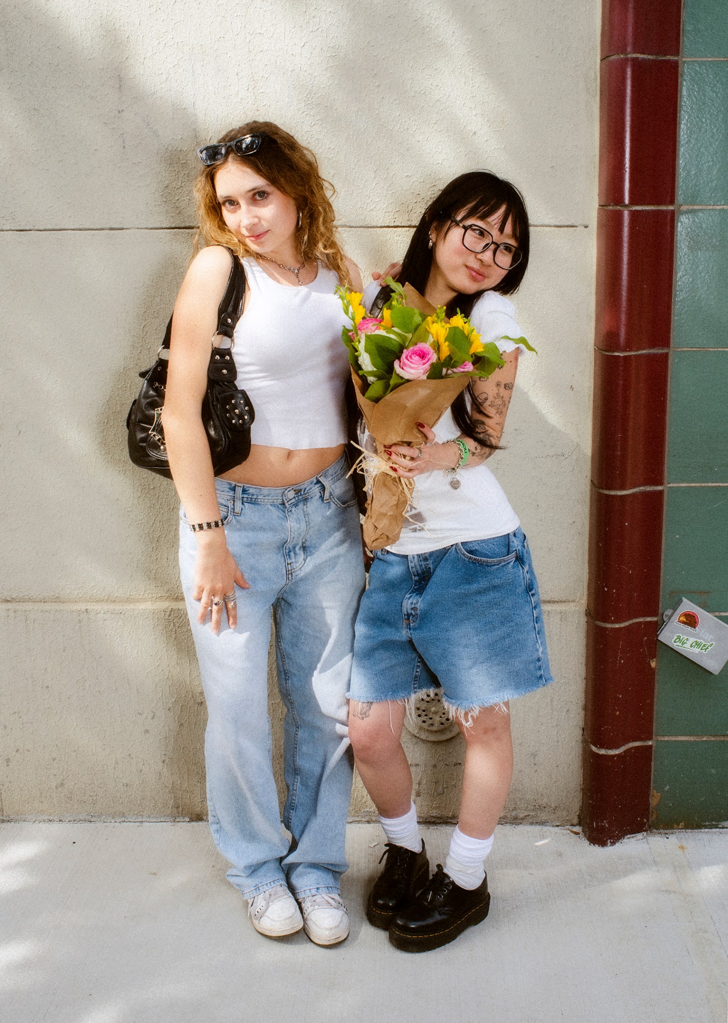 ashley markle photo of a sofia coppola fan on the streets of new york. one wears denim jeans and sneakers and a white vest. they have long blonde hair that's wavy. the other is wearing dnim jorts, black dr martens and is carrying a bunch of flowers