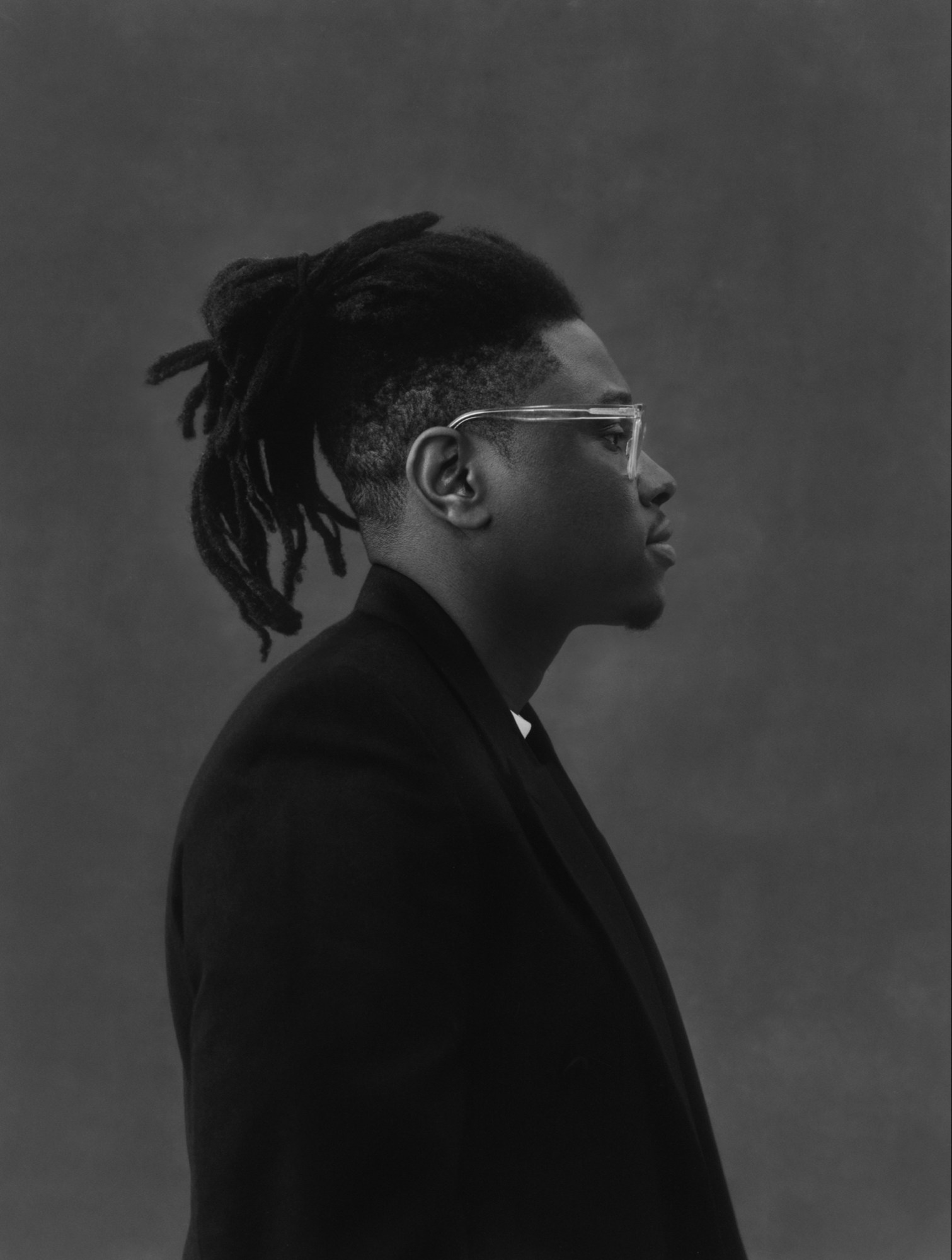 a black & white portrait of the artist Julianknxx sideways on, with clear-framed glasses and his dreads tied up in a ponytail