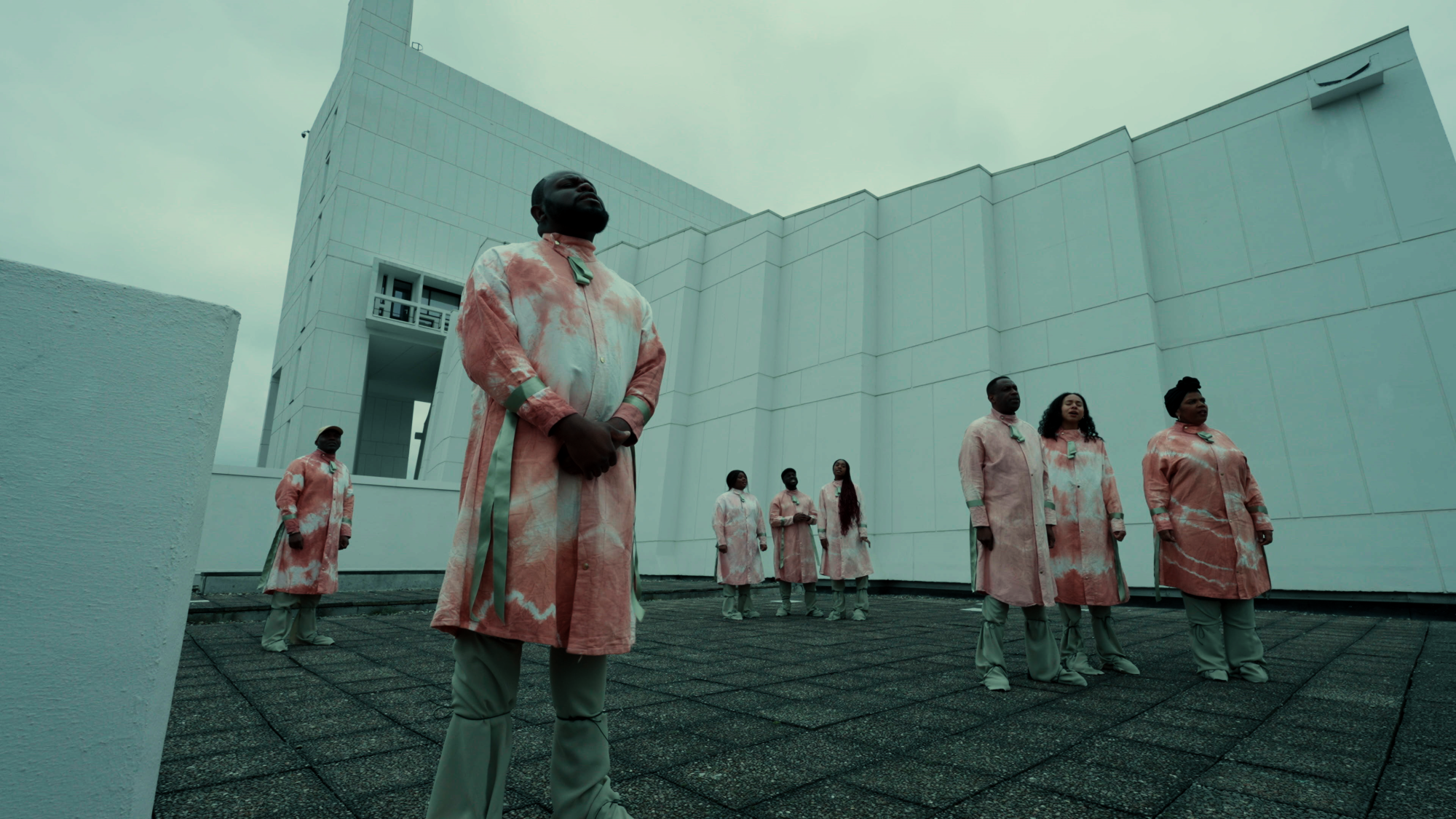 8 people wearing pink and white tie-dyed overshirts stand in small groups on the roof of a building