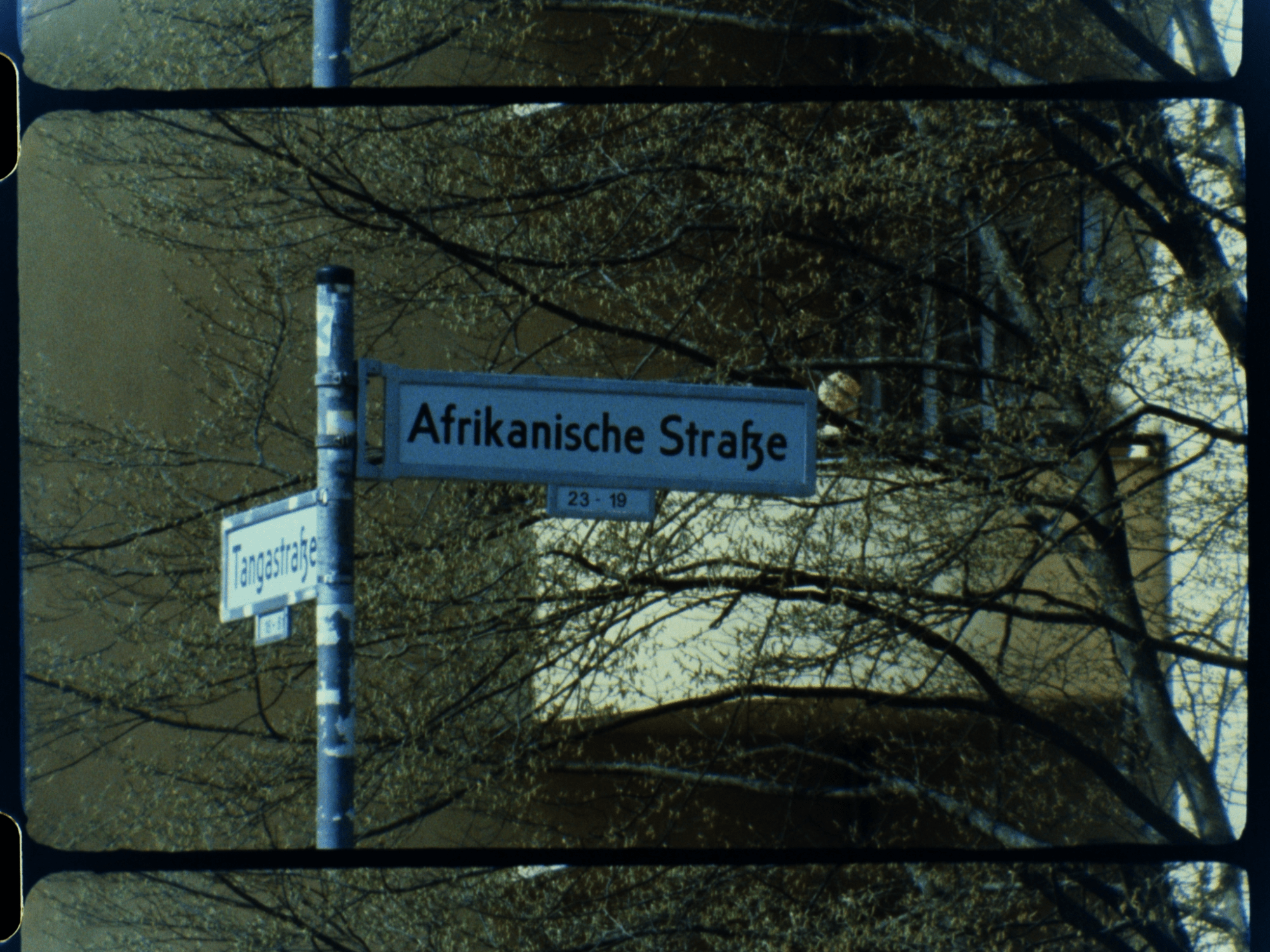 a film still of signposts on a Berlin street corner. one sign reads: Afrikanische Straße