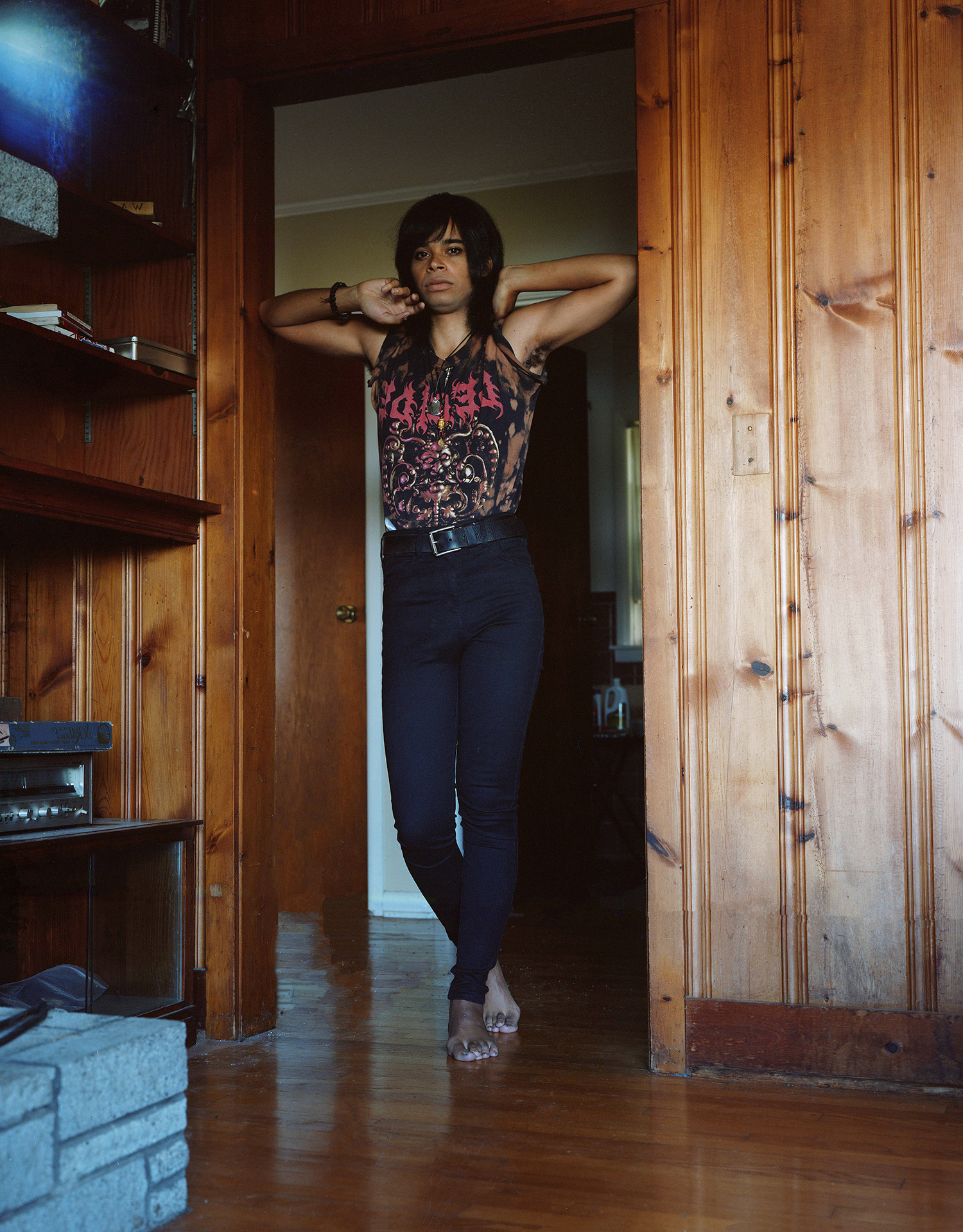 A person stands in a wooden doorframe wearing a black band tee and black skinny jeans