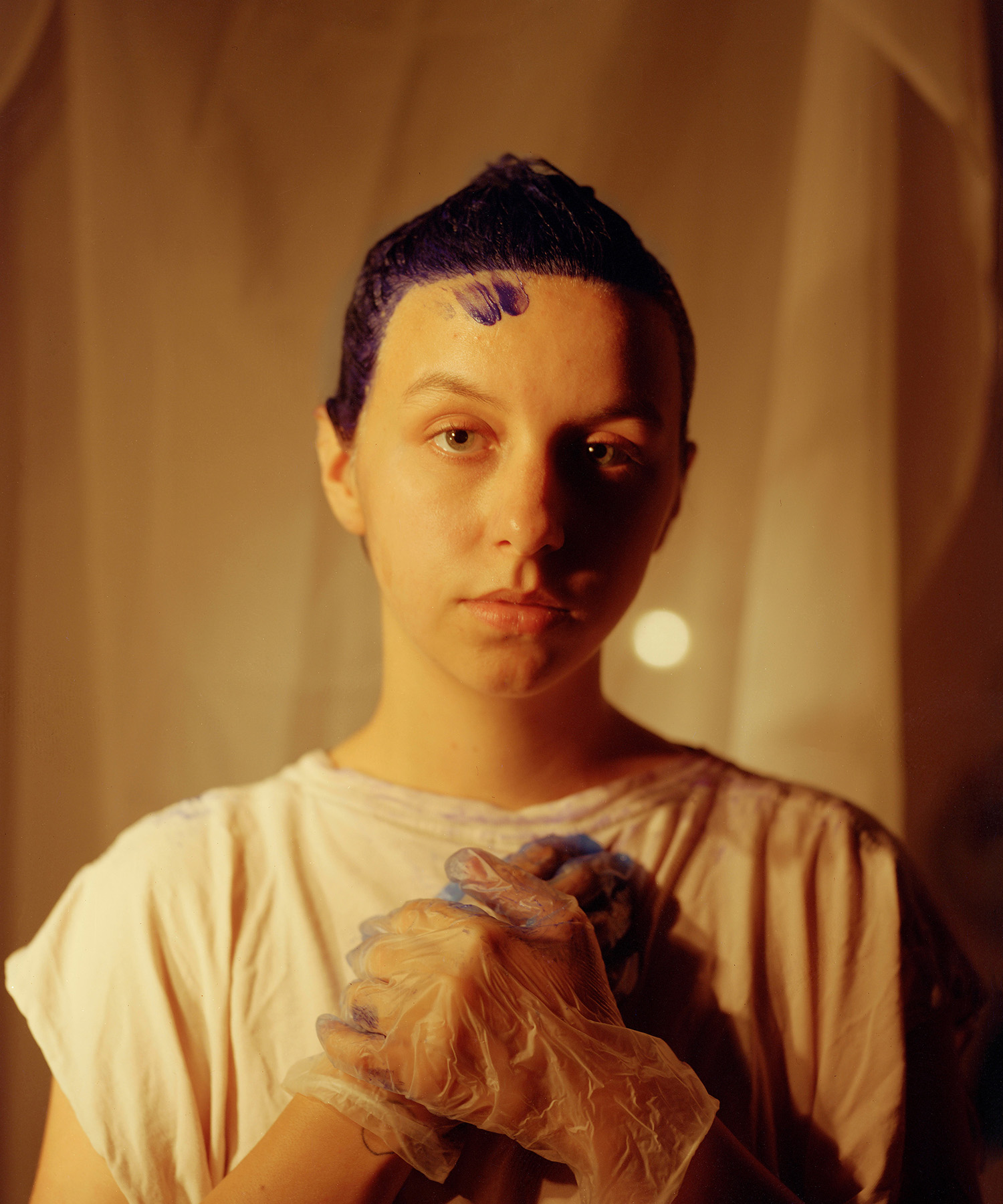 A sepia toned image of a person in white t-shirt with blue hair dye in their hair, wearing disposable gloves also stained blue