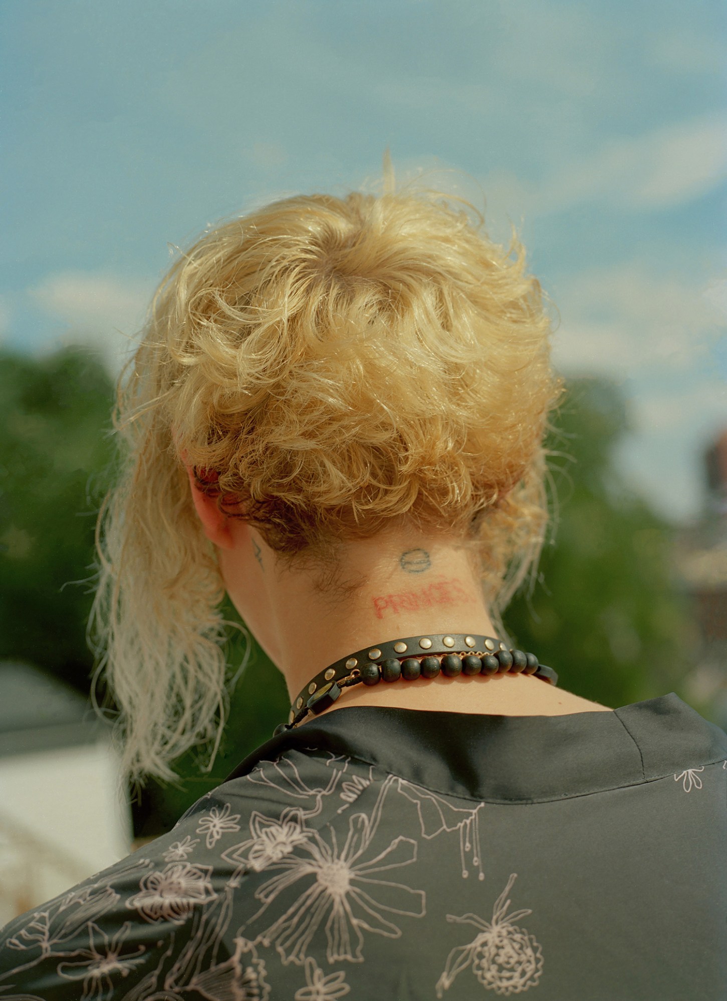 A person with a blonde mullet shot from behind in a black floral shirt, wearing black necklaces