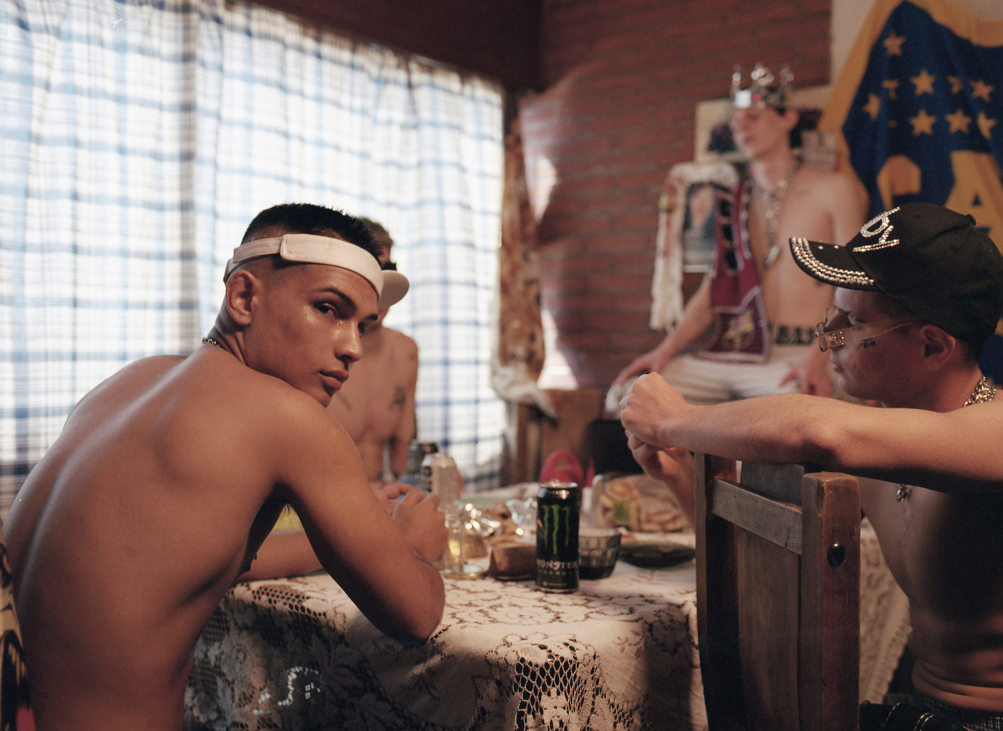 shirtless men in bedazzled hats and visors sit at a kitchen table with a lace tablecloth