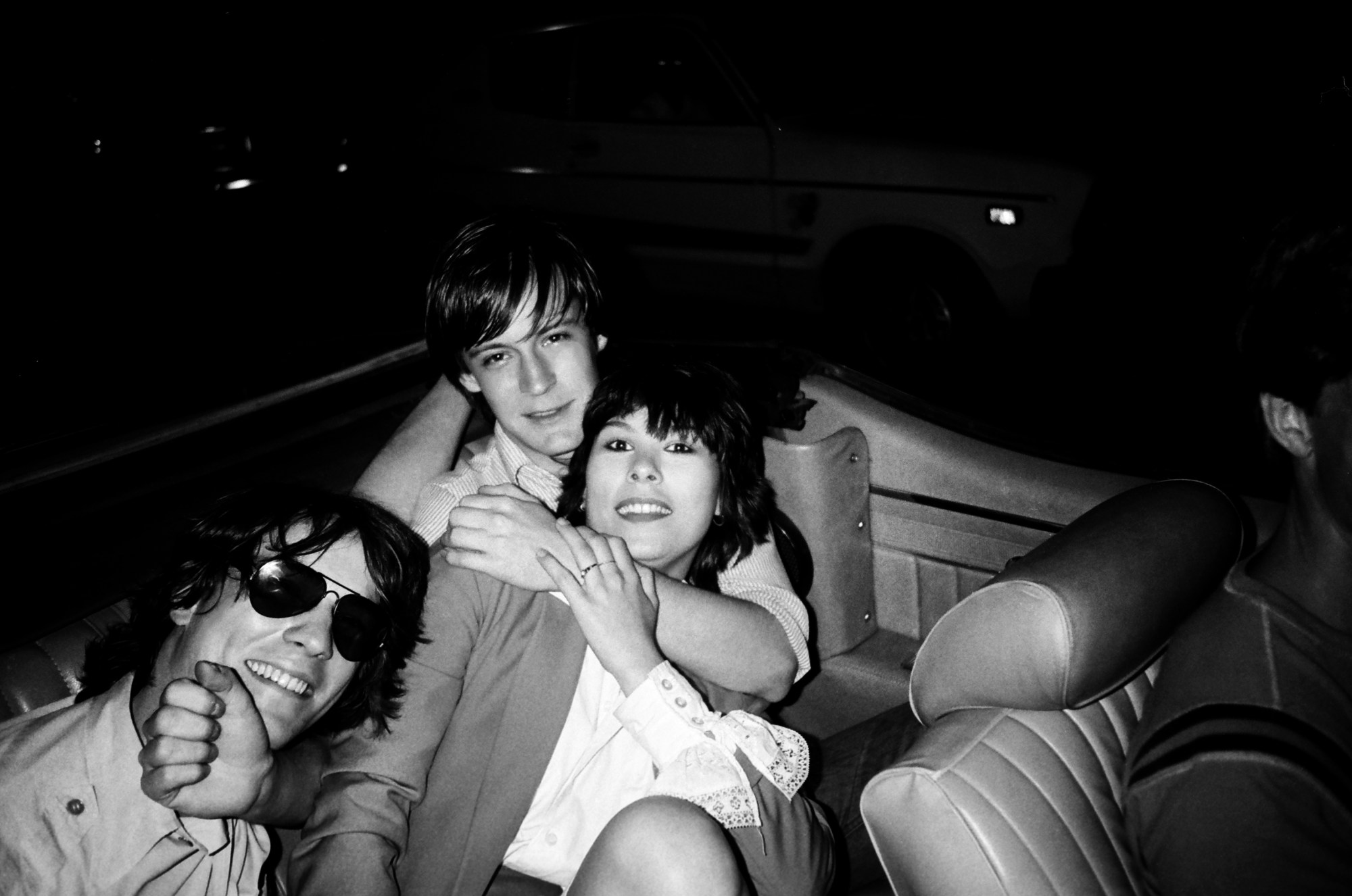 patrick mcmullan photo of three friends in the back of a car in 1980s New York. a man wears sunglasses resting on the shoulder of a woman in a blazer and shirt with dark hair. a man has his hand affectionately round her neck. they all smile.