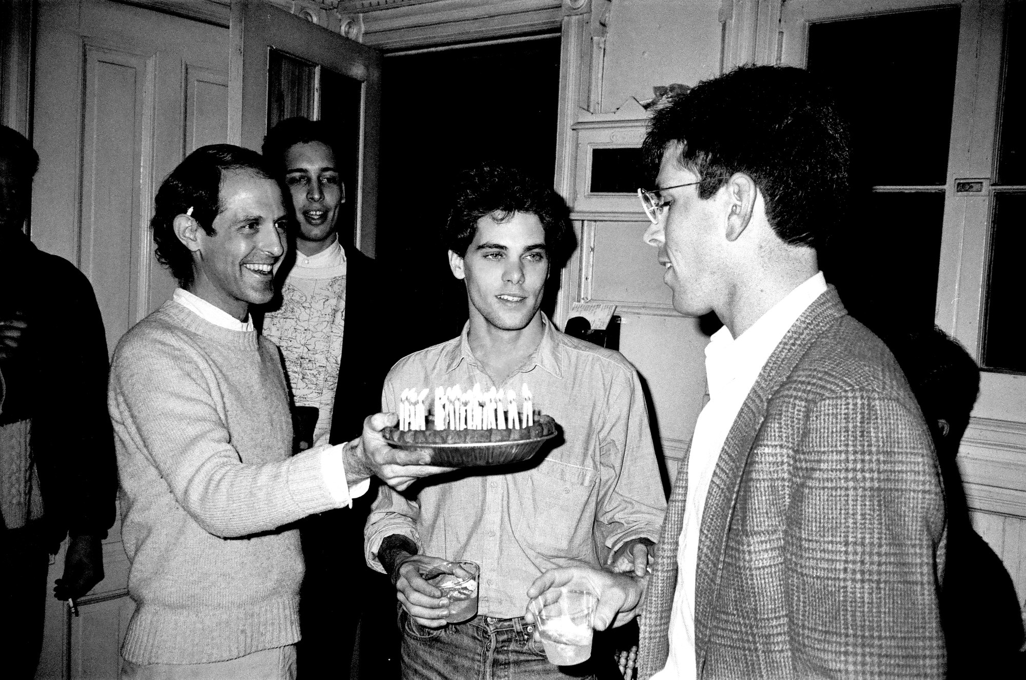 patrick mcmullan photo of chris scott of theater of the ridiculous wearing a sweater holding a cake. scott fritz, a handsome man with curly dark hair stands in the middle wearing a shirt and jeans. jim holt blows out the candles. he is a man wearing glasses and a tartan suit jacket.