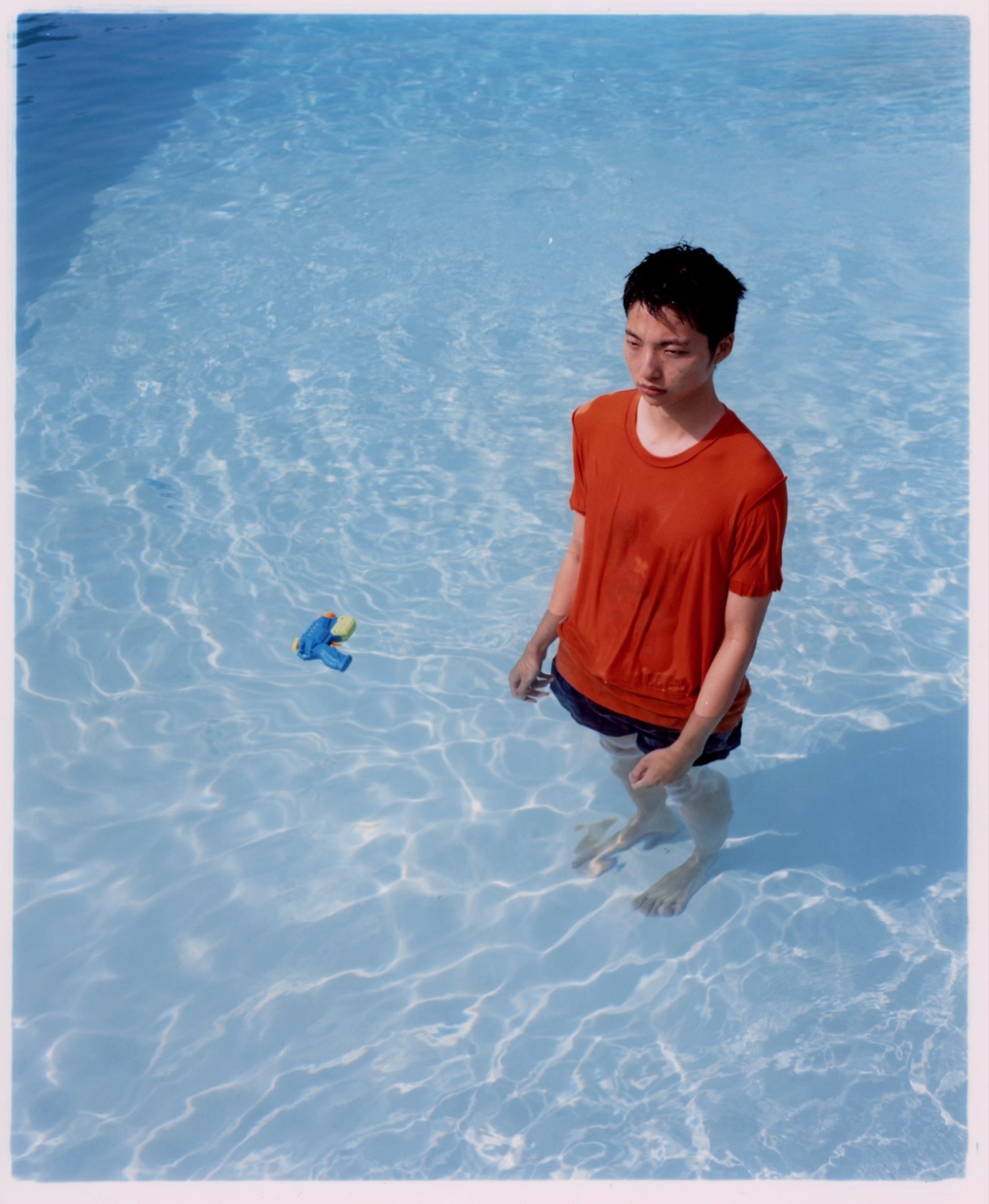 a south east asian man in a wet red t-shirt stands in a swimming pool; a water pistol floats nearby