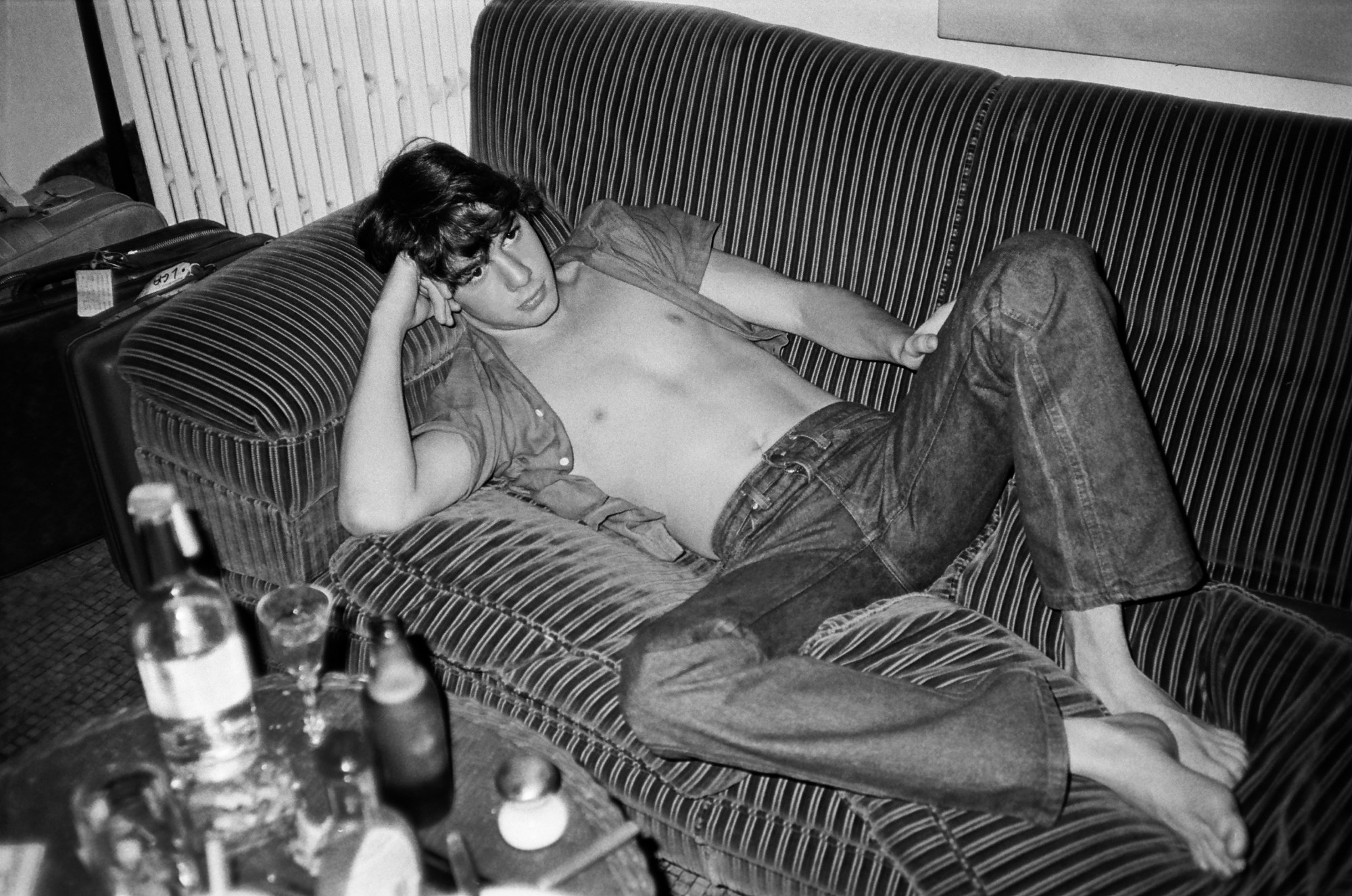 a young man wearing jeans and an open shirt reclines on a stripy sofa; a tabled full of glasses and a vodka bottle cover the coffee table in the foreground