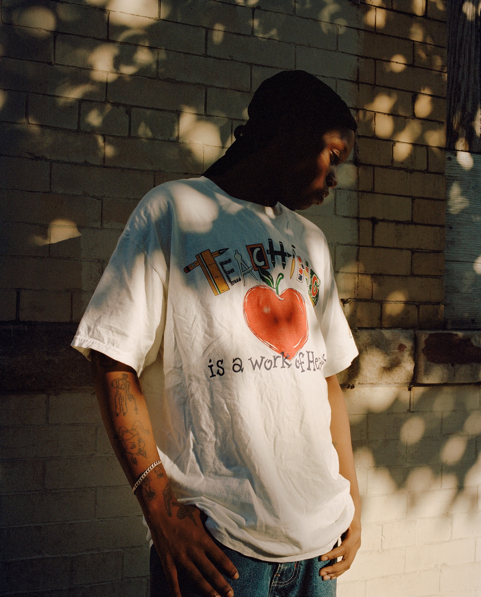 a young Black man with tattoos wearing a novelty vintage T-shirt, standing against a sunny wall