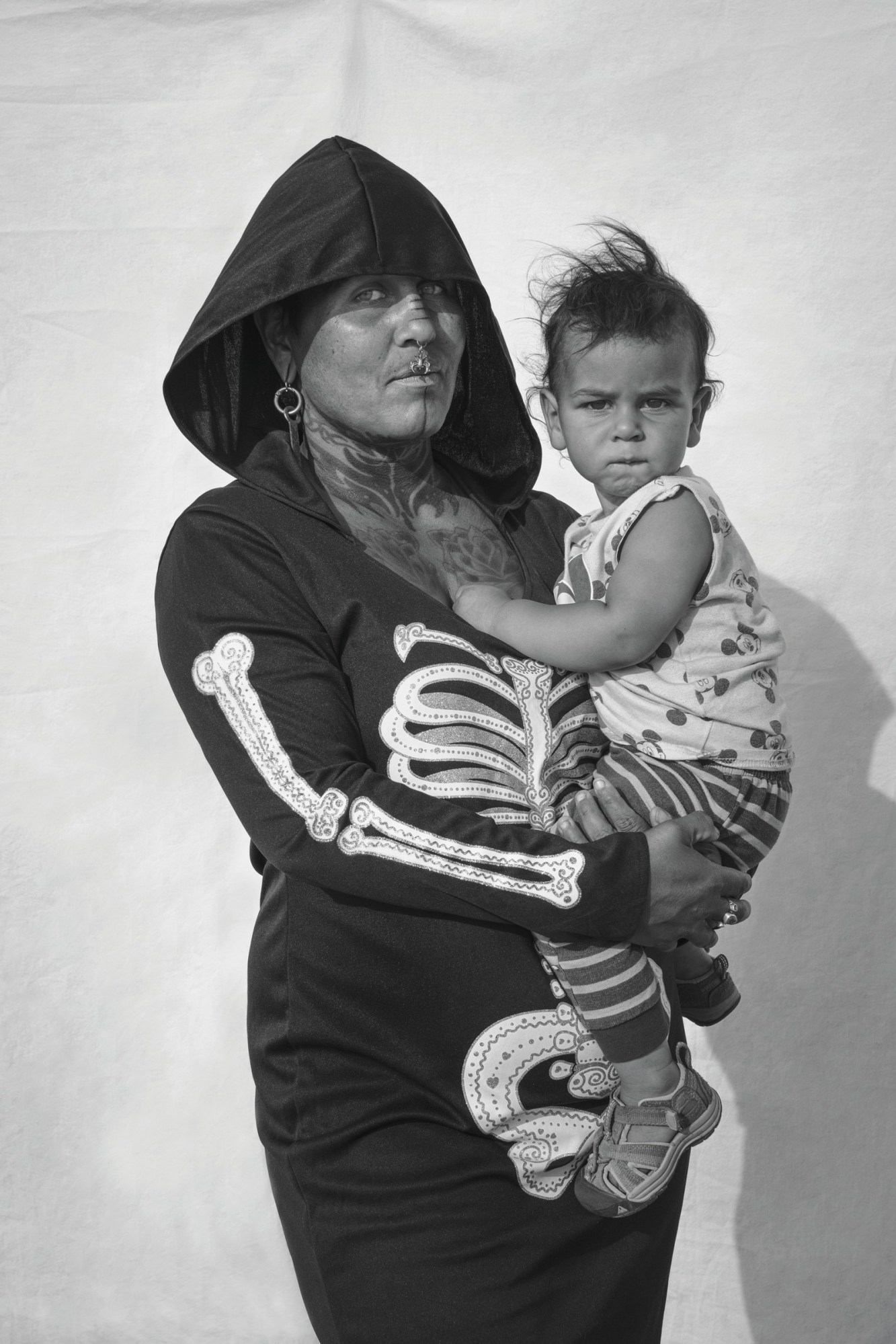 a mother with face tattoos wearing a hooded dress with a skeleton on it, holding her infant child