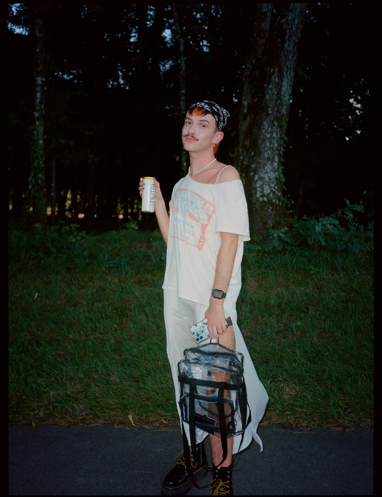 a lana del rey fan stands against grass and trees. they wear doctor marten shoes and a dress, with a budweiser t-shirt over the top of it.