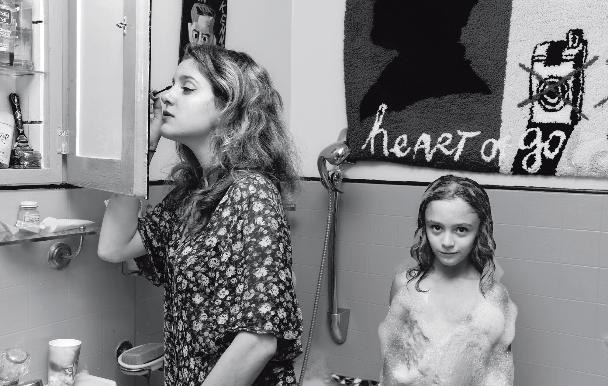 black-and-white image of a teenage girl putting on mascara in bathroom mirror while a younger child stands next to her covered in bubbles