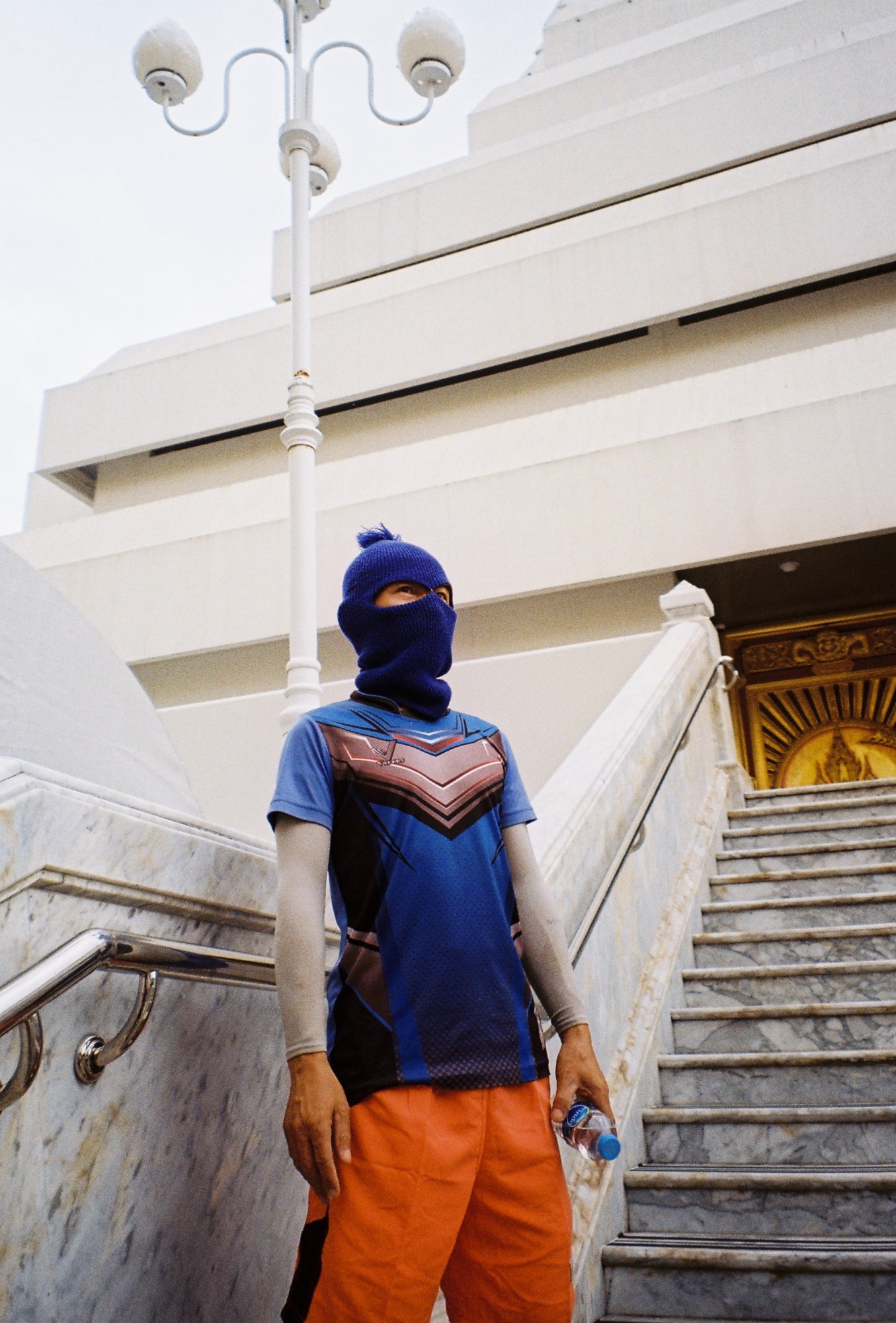 a man in a blue shirt and ski mask wearing orange shorts near a marble staircase