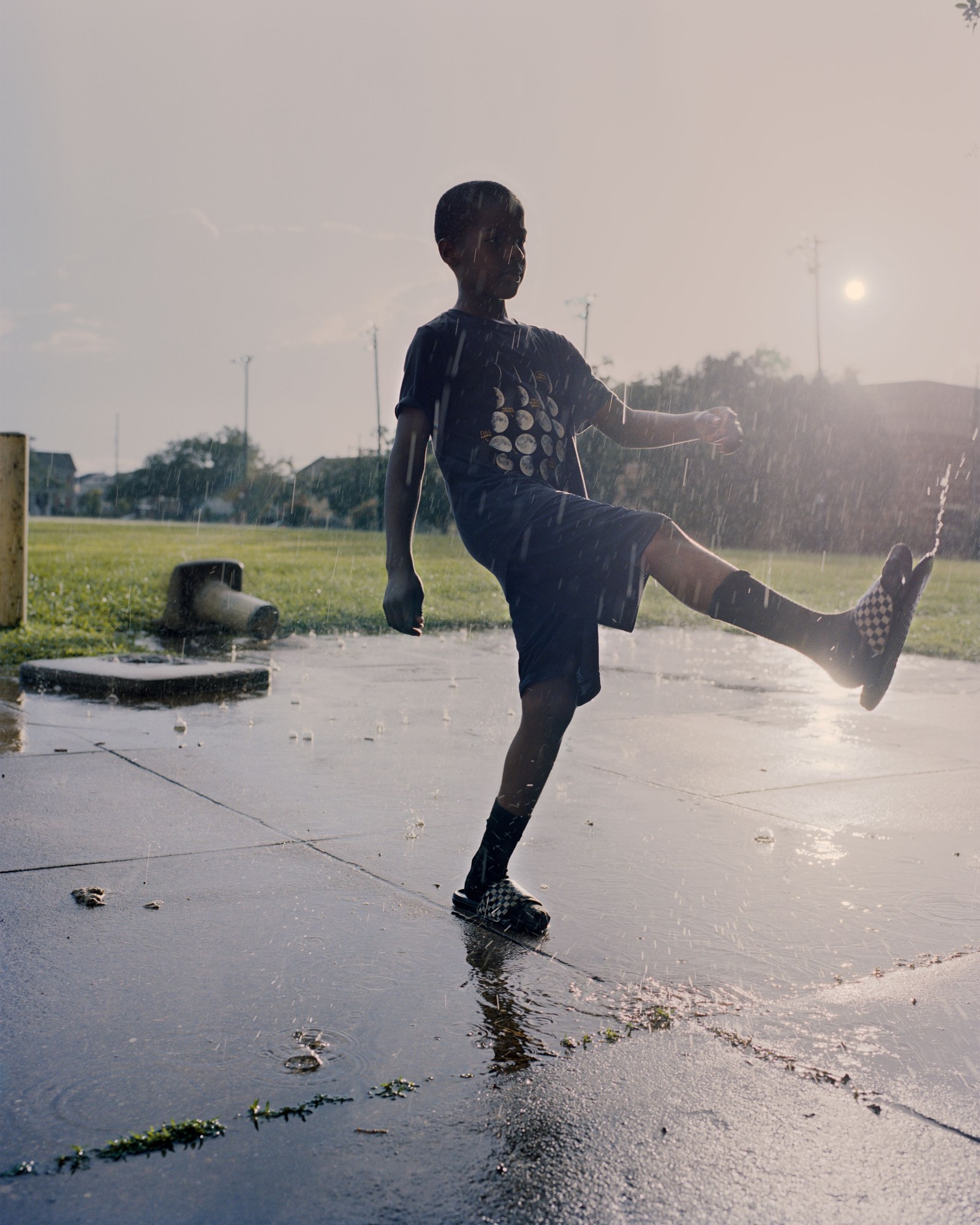 Photograph by Vasantha Yogananthan from Mystery Streets of a boy kicking rain puddles in a playground