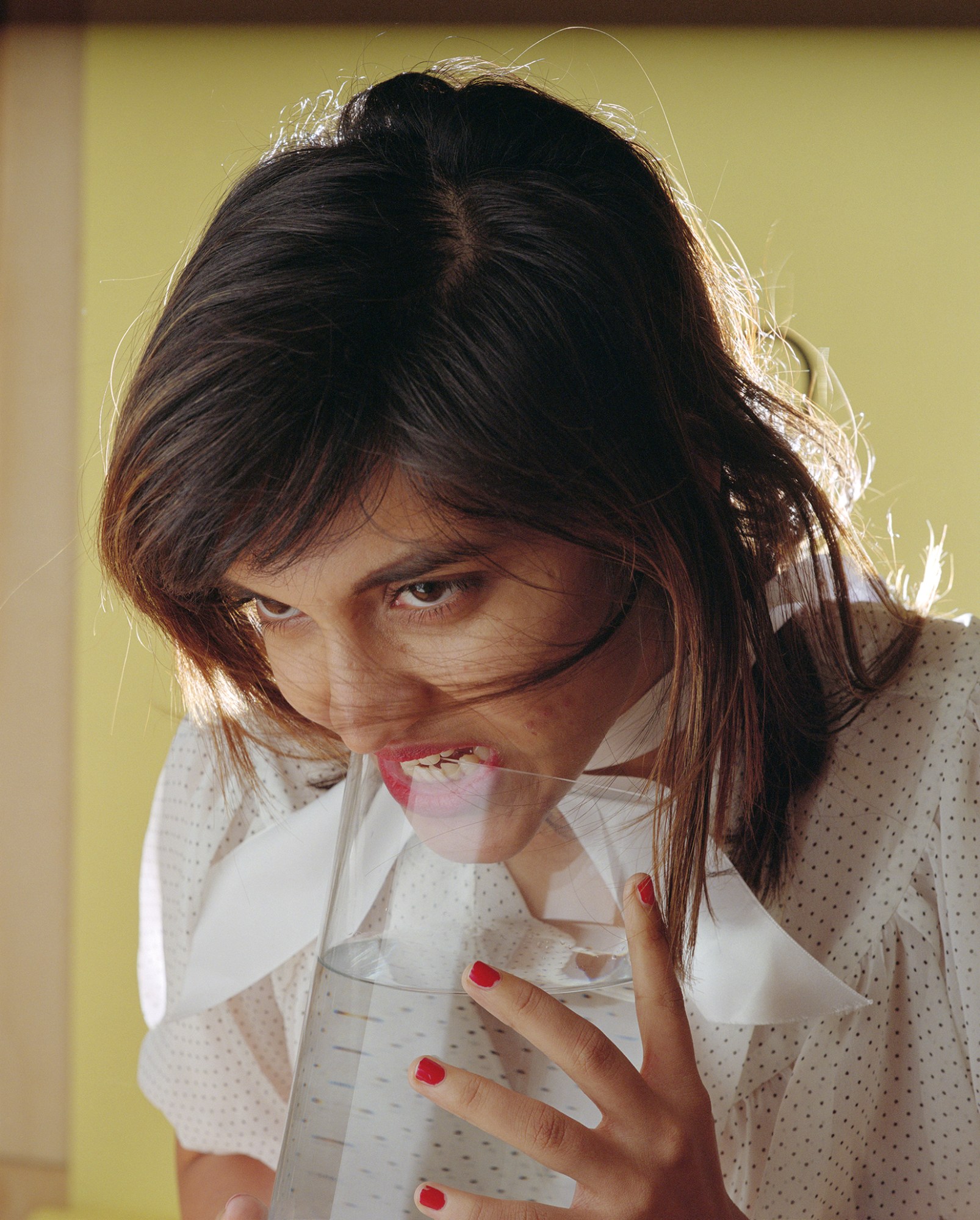 a young brunette woman in a polkadot dress bites the top of an oversized glass of water