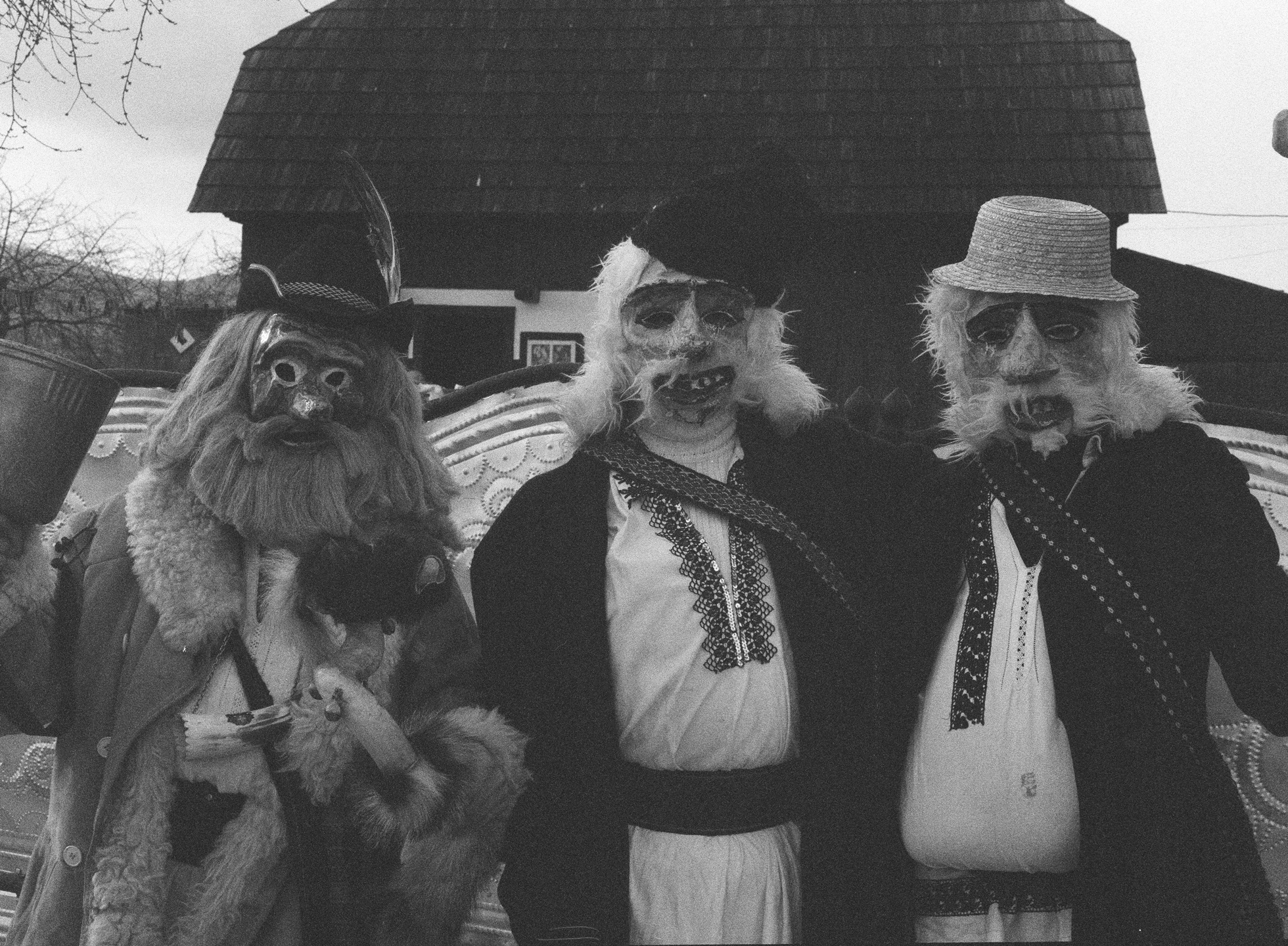 a group of three people wearing old man or gnome costumes and papiere mache-like masks stand together outside a house