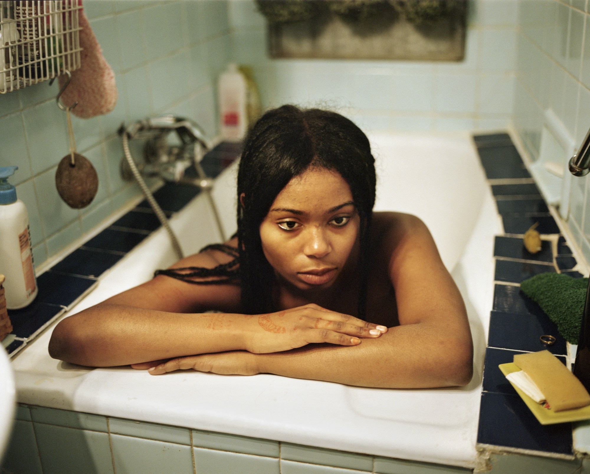 a young Black woman with straight hair sits in a bathtub in a messy, dated bathroom