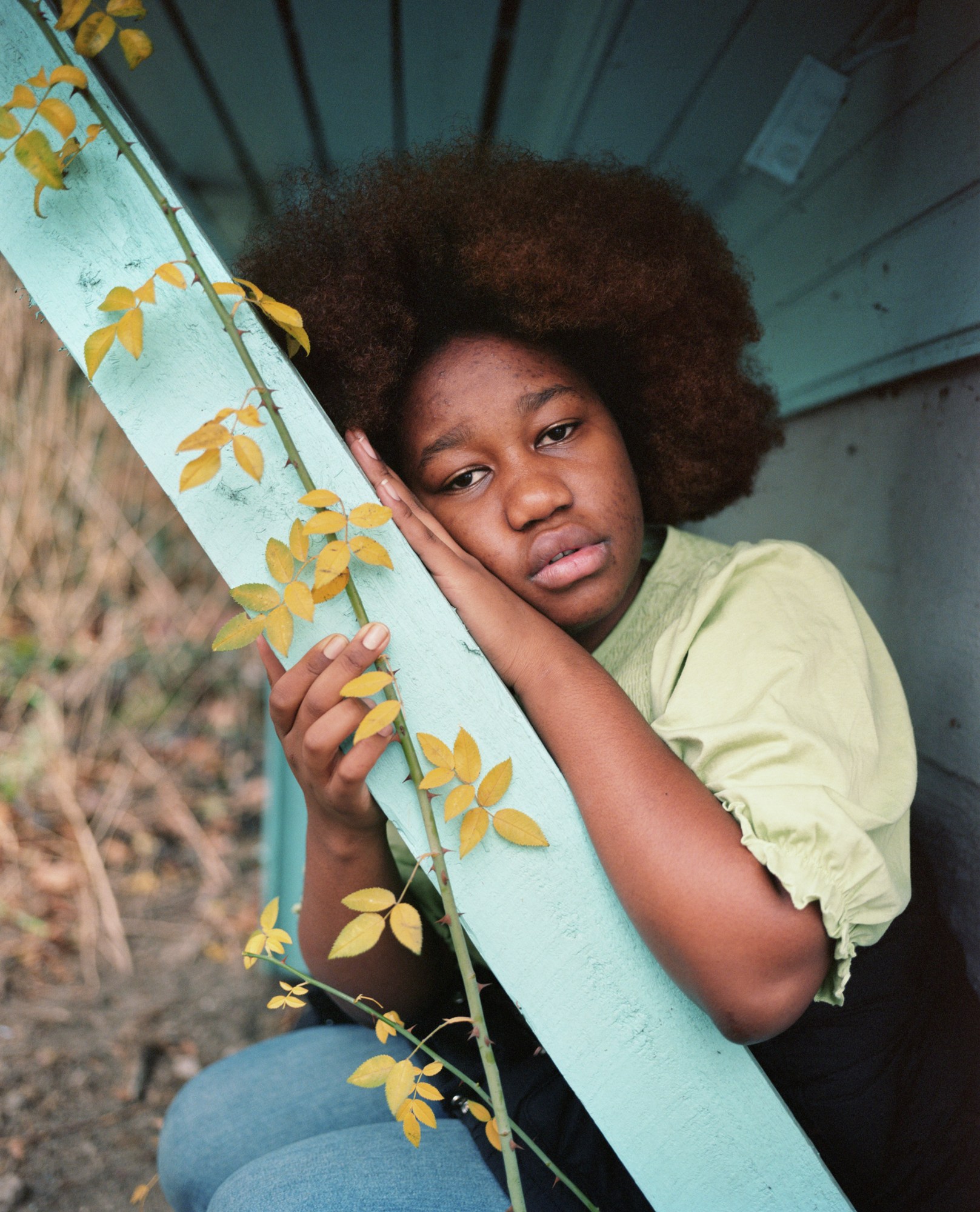 a young Black woman with an afro wears jeans and a pale green blouse; she sits under a wooden shelter, leaning on a beam