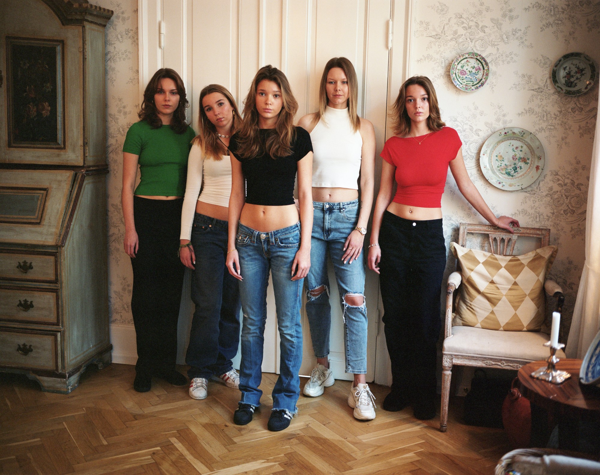 five teenage white girls wearing jeans and crop tops stand in a living room full of antique furniture