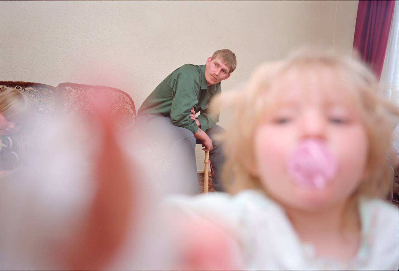 Photograph by Nick Waplington of a man sat in a living room with a kid with a dummy playing with the camera and another on the sofa from The Living Room Series.