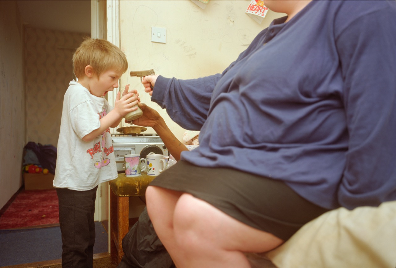 Photograph by Nick Waplington of a woman sat pointing a toy gun at a child head who is holding a mug, taken from the book ‘Weddings, Parties, Anything’