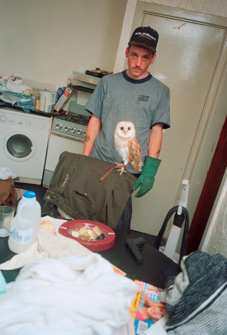 Photograph by Nick Waplington of a man with an owl standing in a dirty kitchen from the book