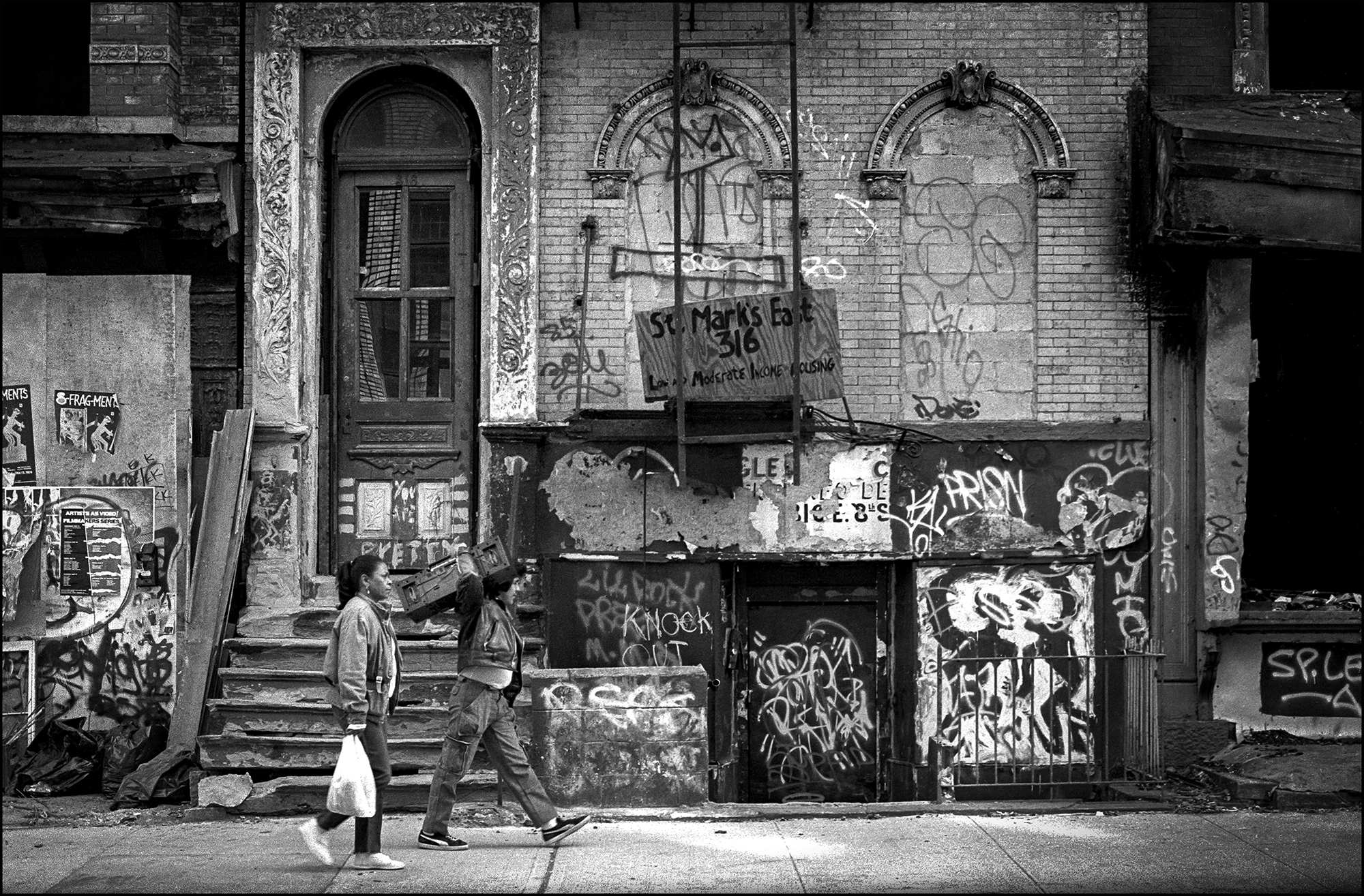 A young man carries an oversized ghetto blaster on his shoulder, passing the deteriorated and graffiti-covered tenement blocks of the Lower East Side.