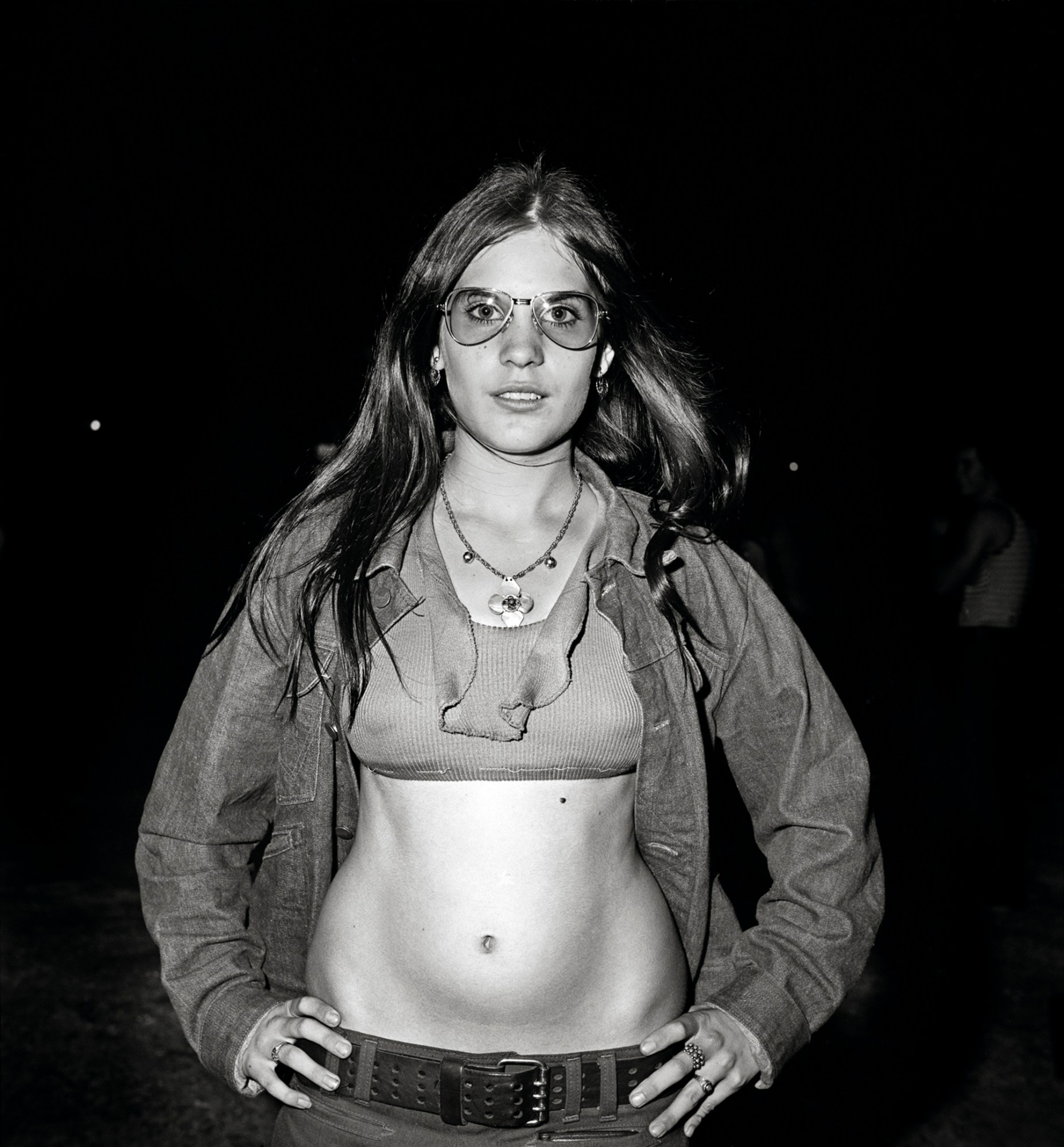 a teenage girl in the 1970s wearing aviator glasses and a crop top with a denim jacket