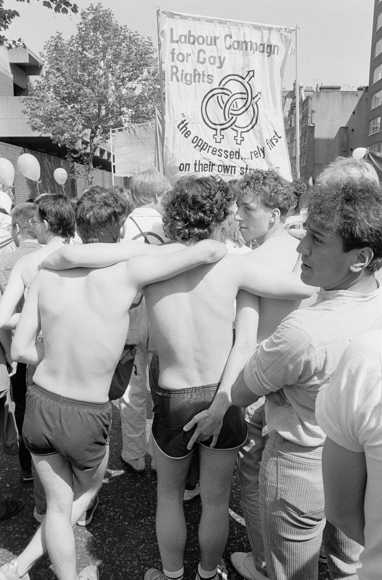 a parade of queer protestors walk shirtless away from the camera, arms round each other. one man has his hand on his friend's buttocks