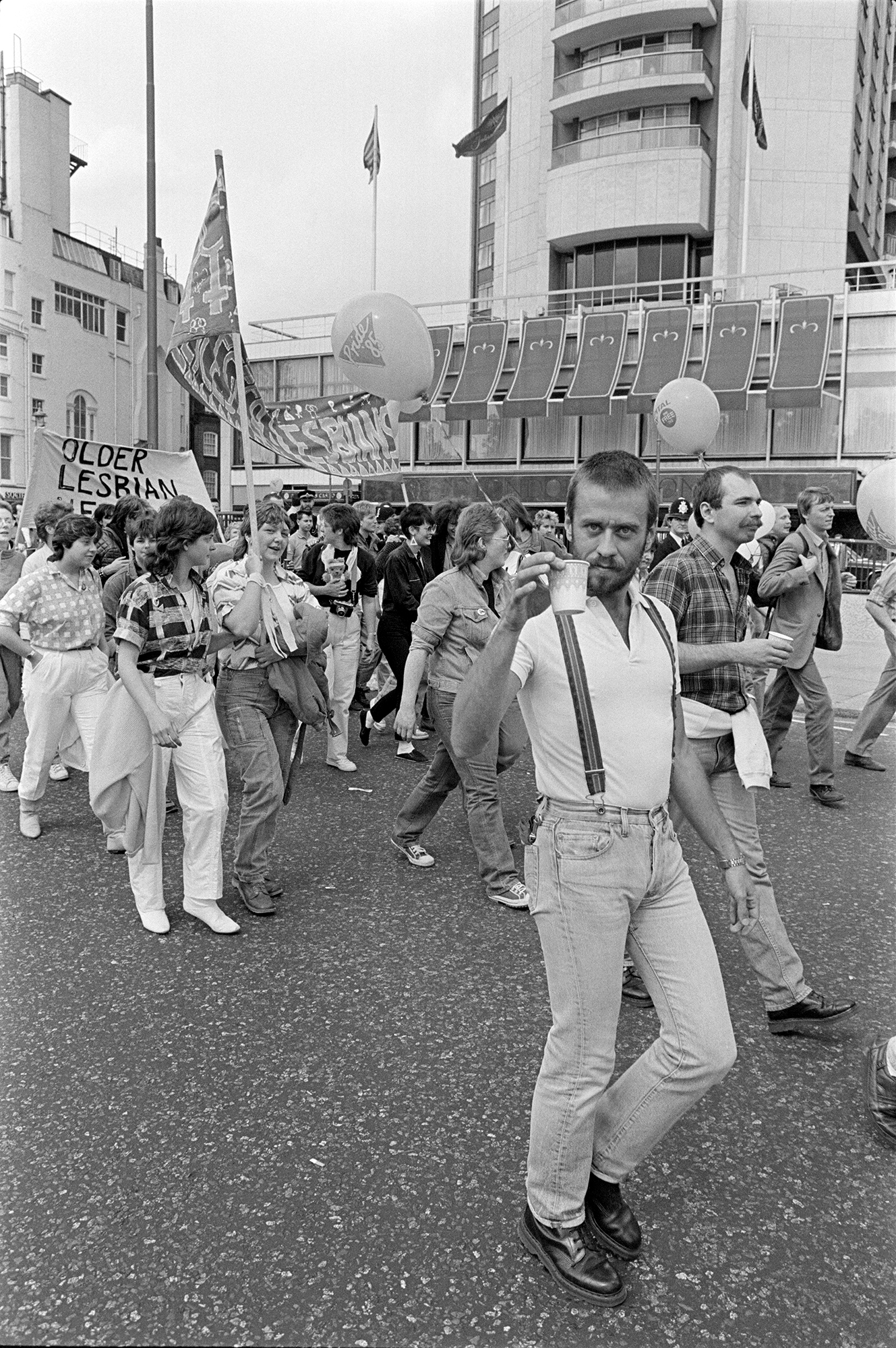 a gay rights protest in central london in the 1980s, a man in suspenders and a white shirt walks next to the camera and looks at it while holding up a cup