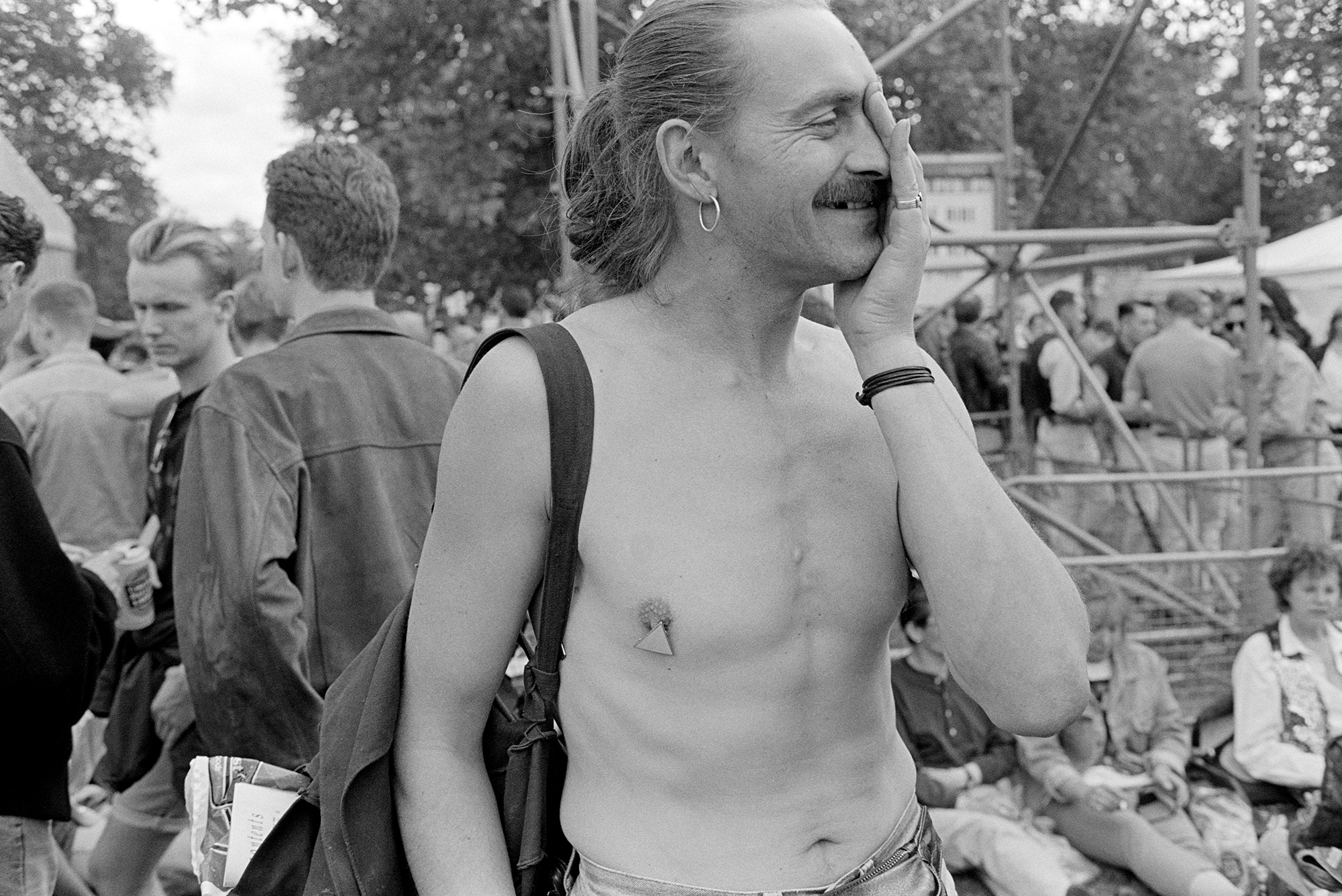 a queer protestor with triangles on his nipples rubs his eye. he has long hair and a moustache and an earring