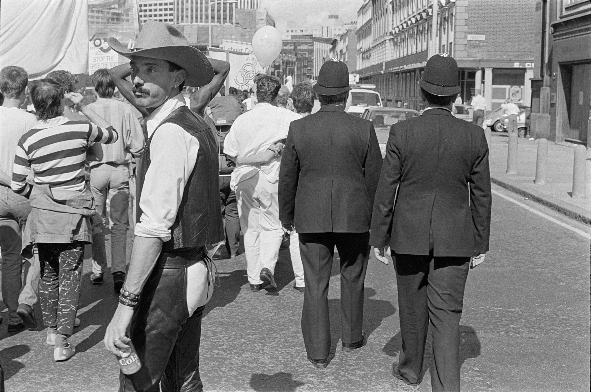 a man dressed as a cowboy with hat and chaps looks off camera as a protest passes by. police offers in uniform follow it, their backs to the camera